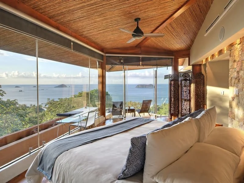 A bedroom with large glass windows overlooking the ocean, with outdoor chairs and a small table on a balcony, and wooden ceiling with a ceiling fan.