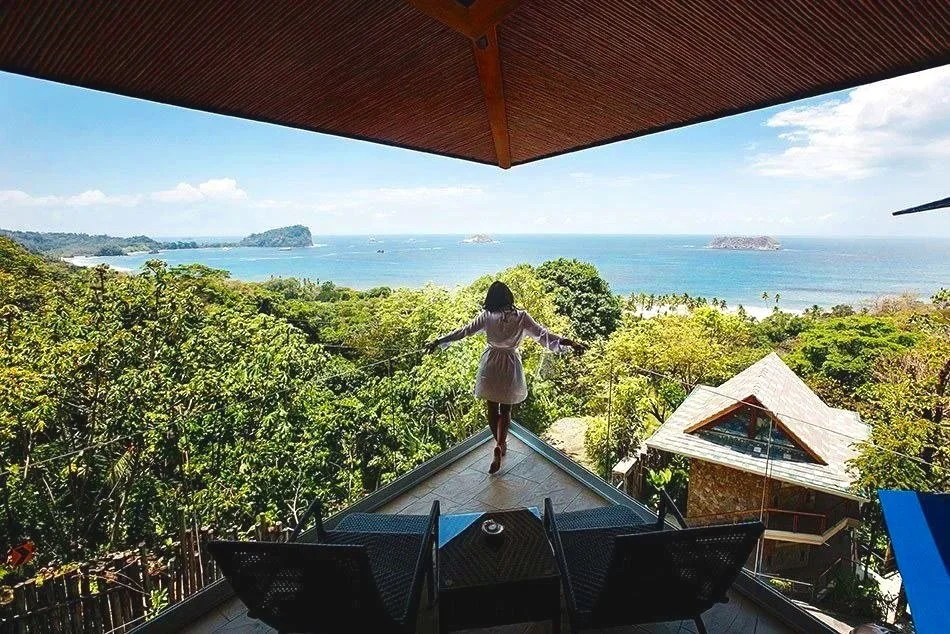 A woman in a white dress standing on a balcony, looking at a lush green landscape and the ocean with small islands in the distance.