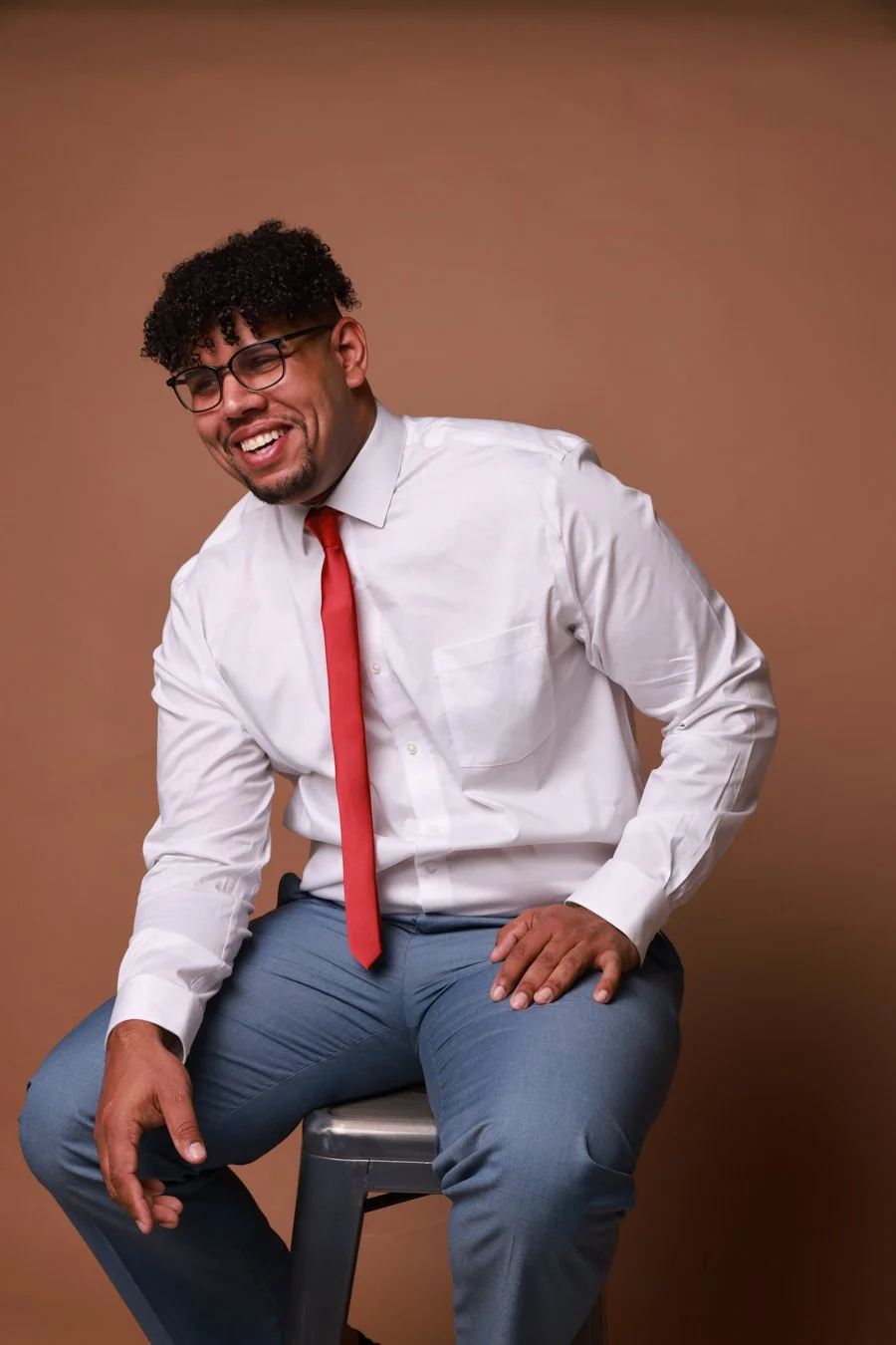 A smiling man with curly hair and glasses wearing a white shirt, red tie, and gray pants, sitting on a stool against a plain brown background.