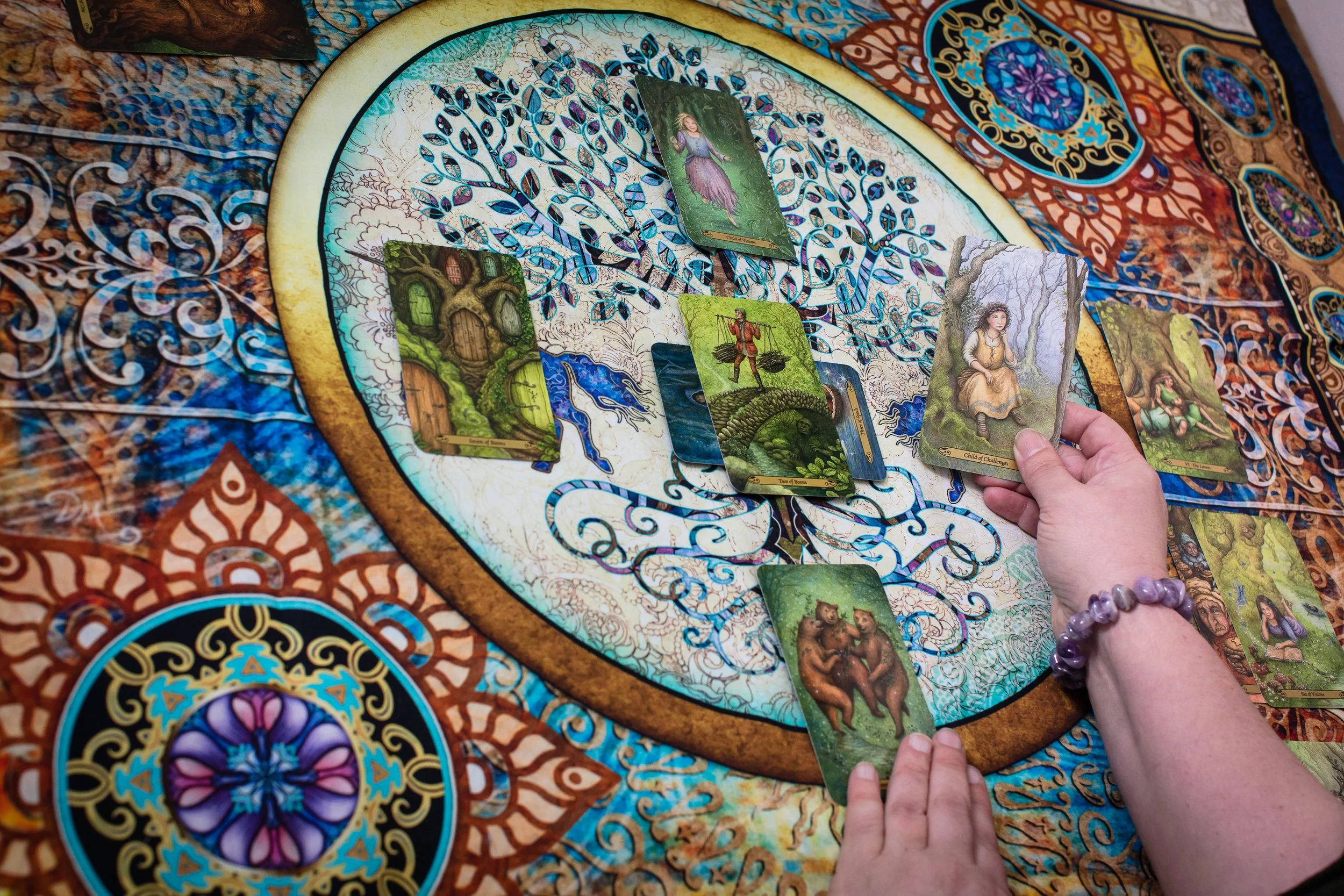 A person is performing a tarot card reading on a colorful, decorated cloth with a circular design. The person holds a card featuring a woman in a yellow dress, while other tarot cards with various mystical and fantasy-themed illustrations are laid out on the cloth and held in their hands.