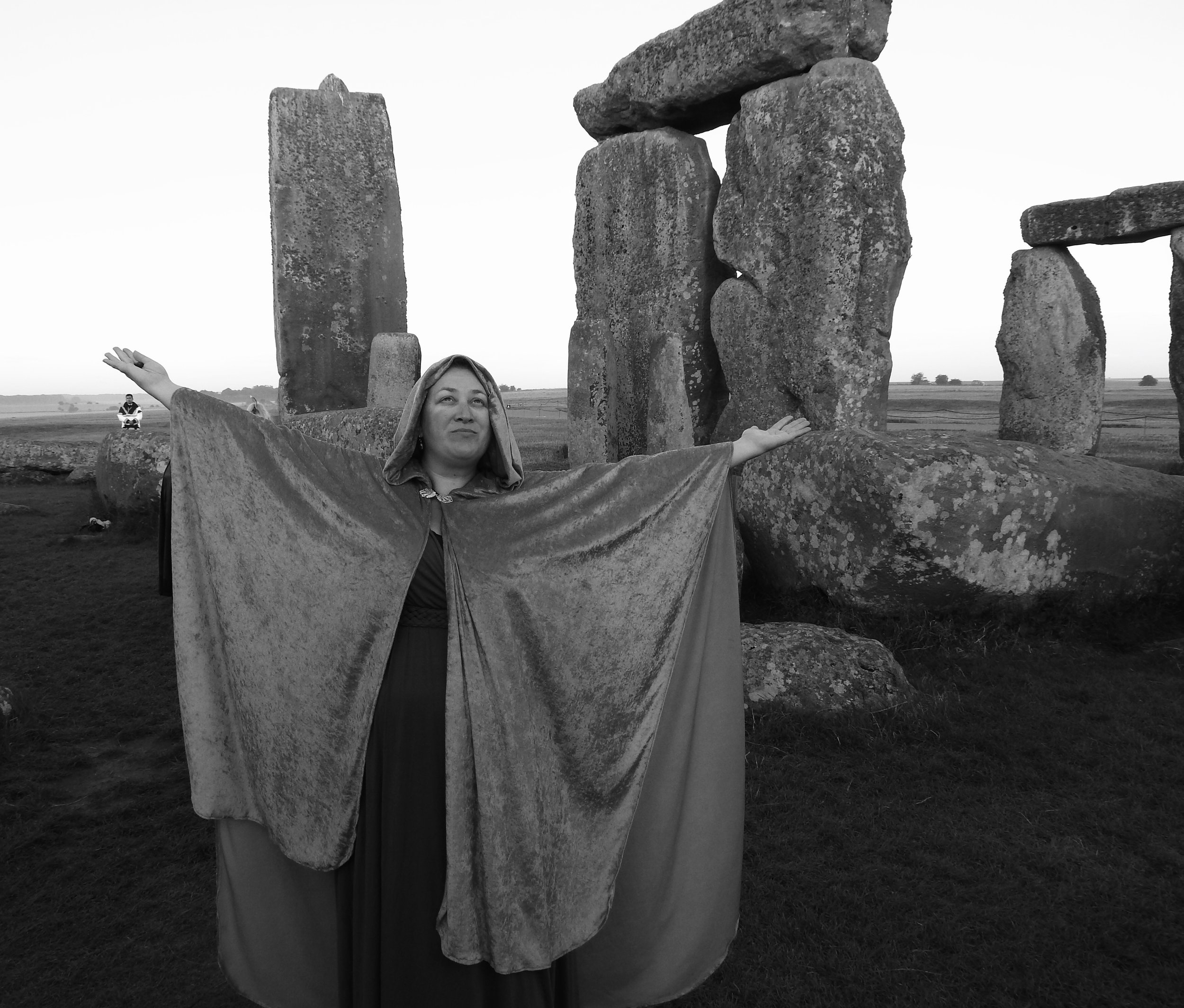 A woman dressed as a medieval or fantasy character with arms outstretched stands in front of Stonehenge, a prehistoric monument in England, during the daytime.
