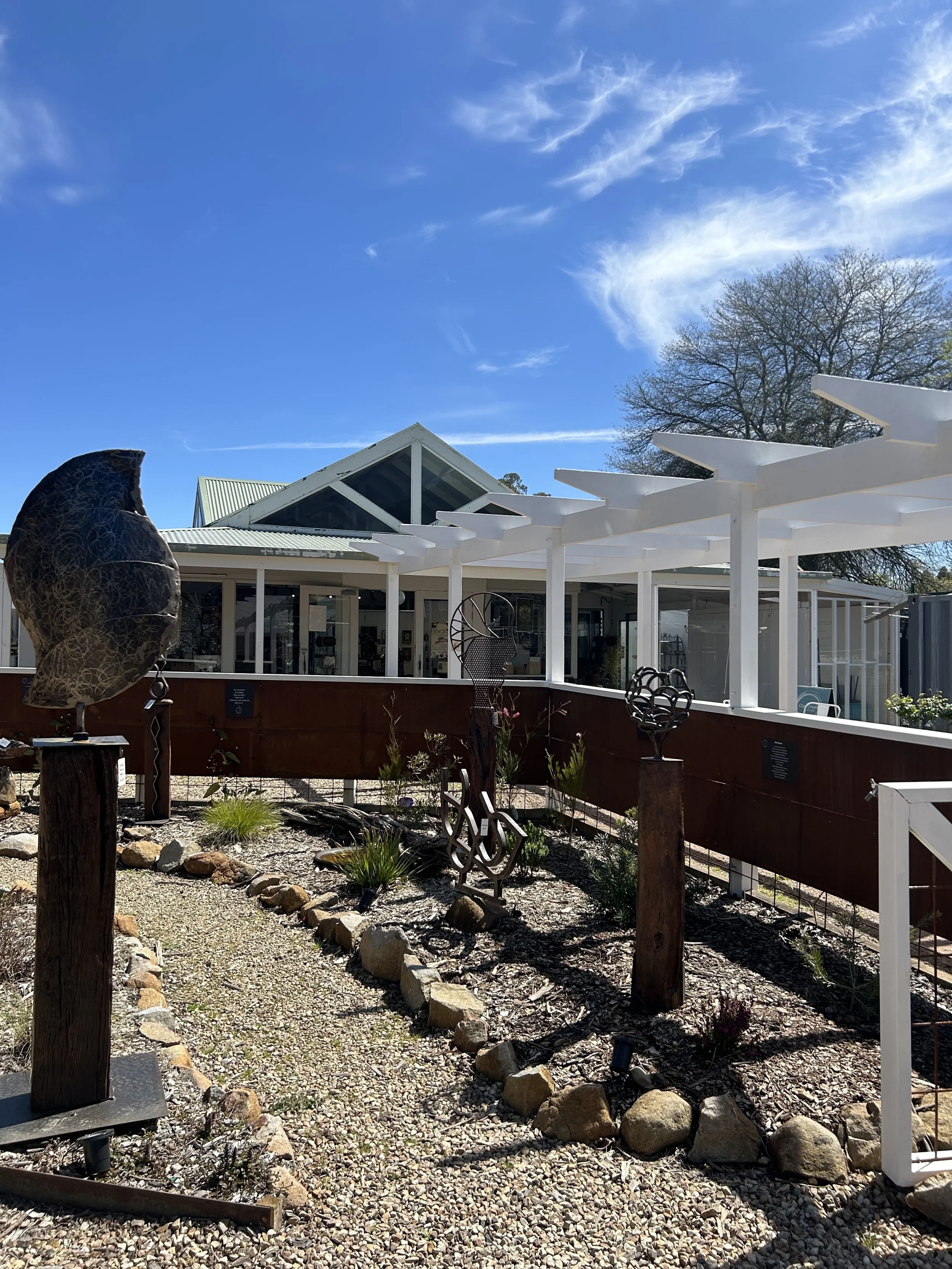 Outdoor area with sculptures, gravel pathway, and modern white building with glass and metal roof under a blue sky with wispy clouds.