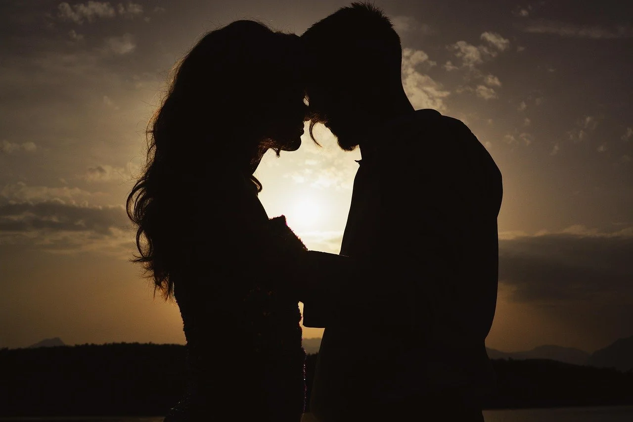 Silhouettes of a man and woman standing close together, touching foreheads, during sunset.