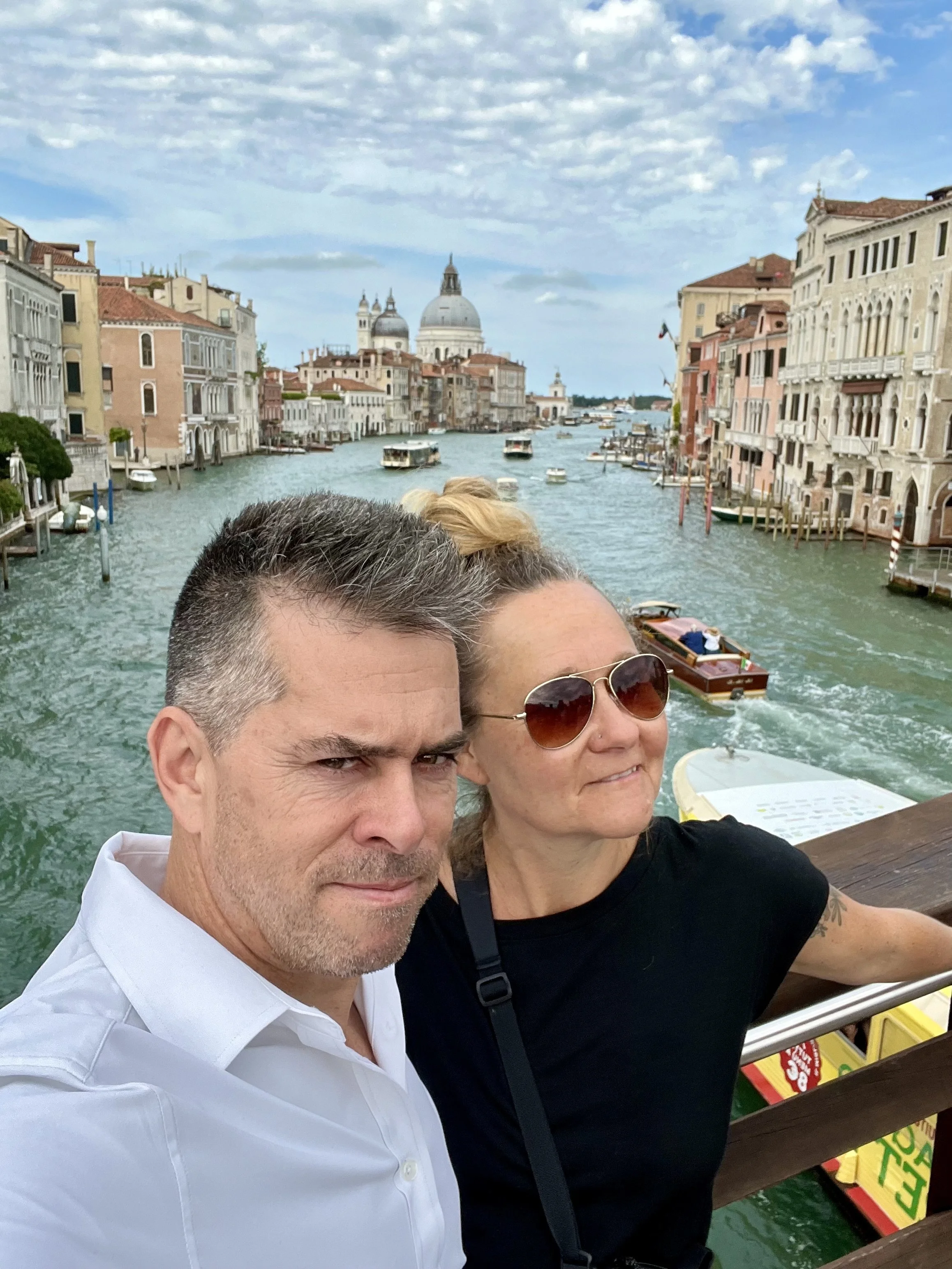 A selfie of a man and a woman on a boat in Venice, Italy, with the Grand Canal and historic buildings in the background.