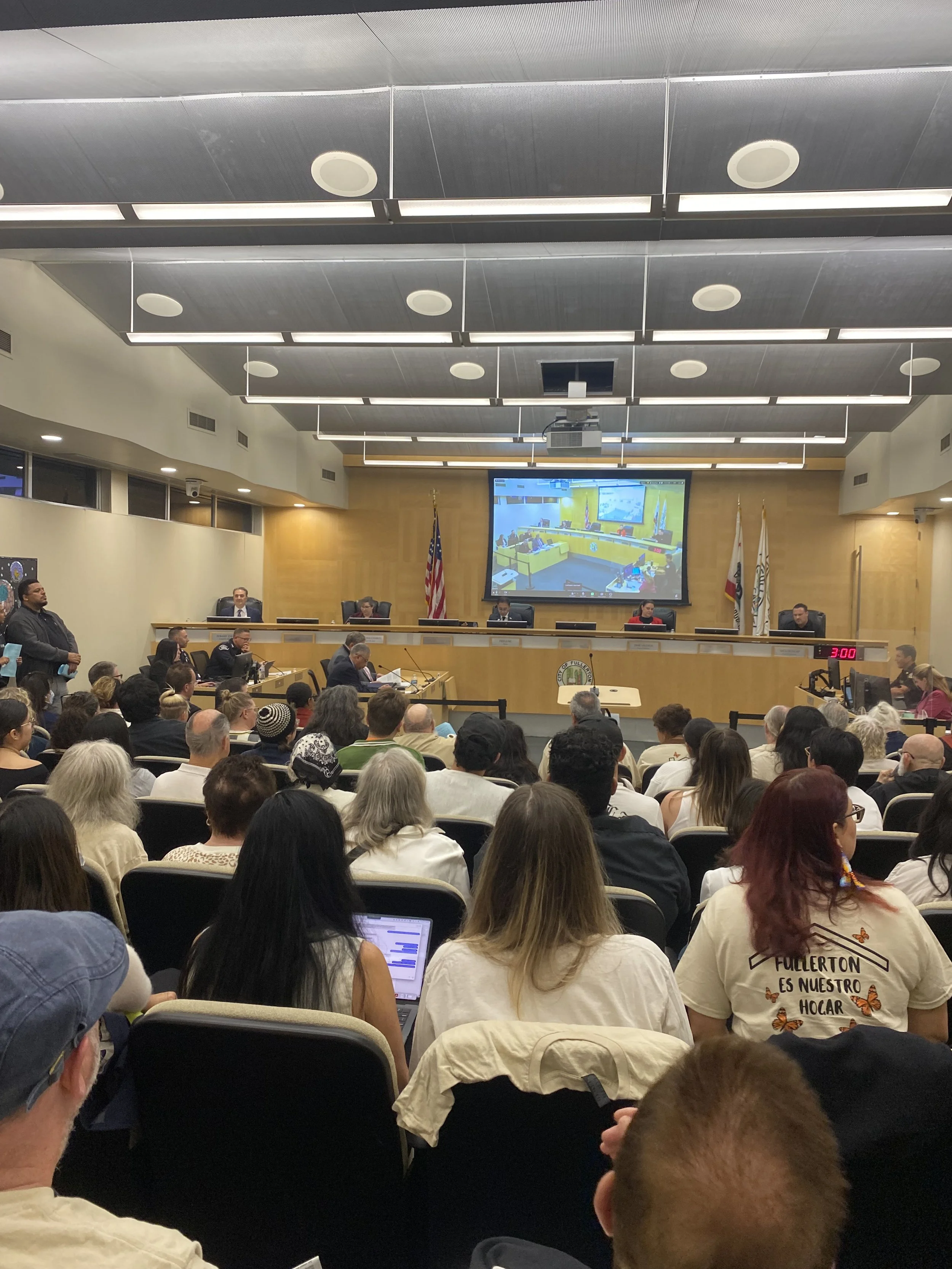 A crowded city council or government meeting with officials seated on a stage in a large conference room. There is a large screen displaying a live video feed of the room, the American flag and other flags are visible behind the officials, and people are attending the meeting with some using laptops and others taking notes.
