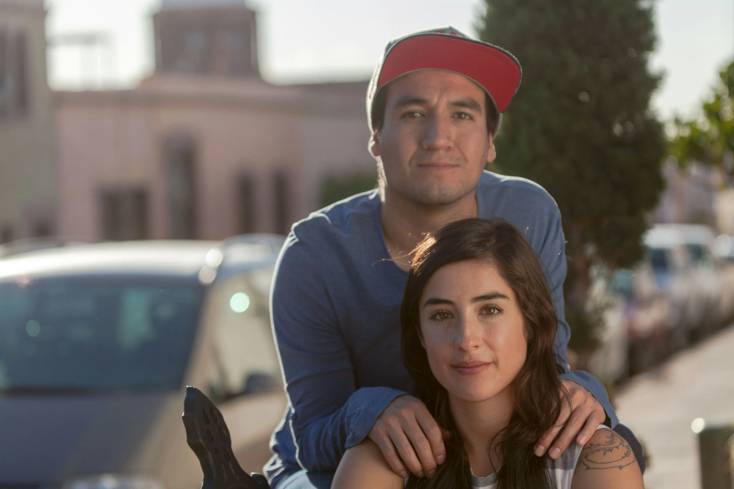 Joven pareja en una calle, con la mujer en primer plano y el hombre detrás, ambos mirando a la cámara.