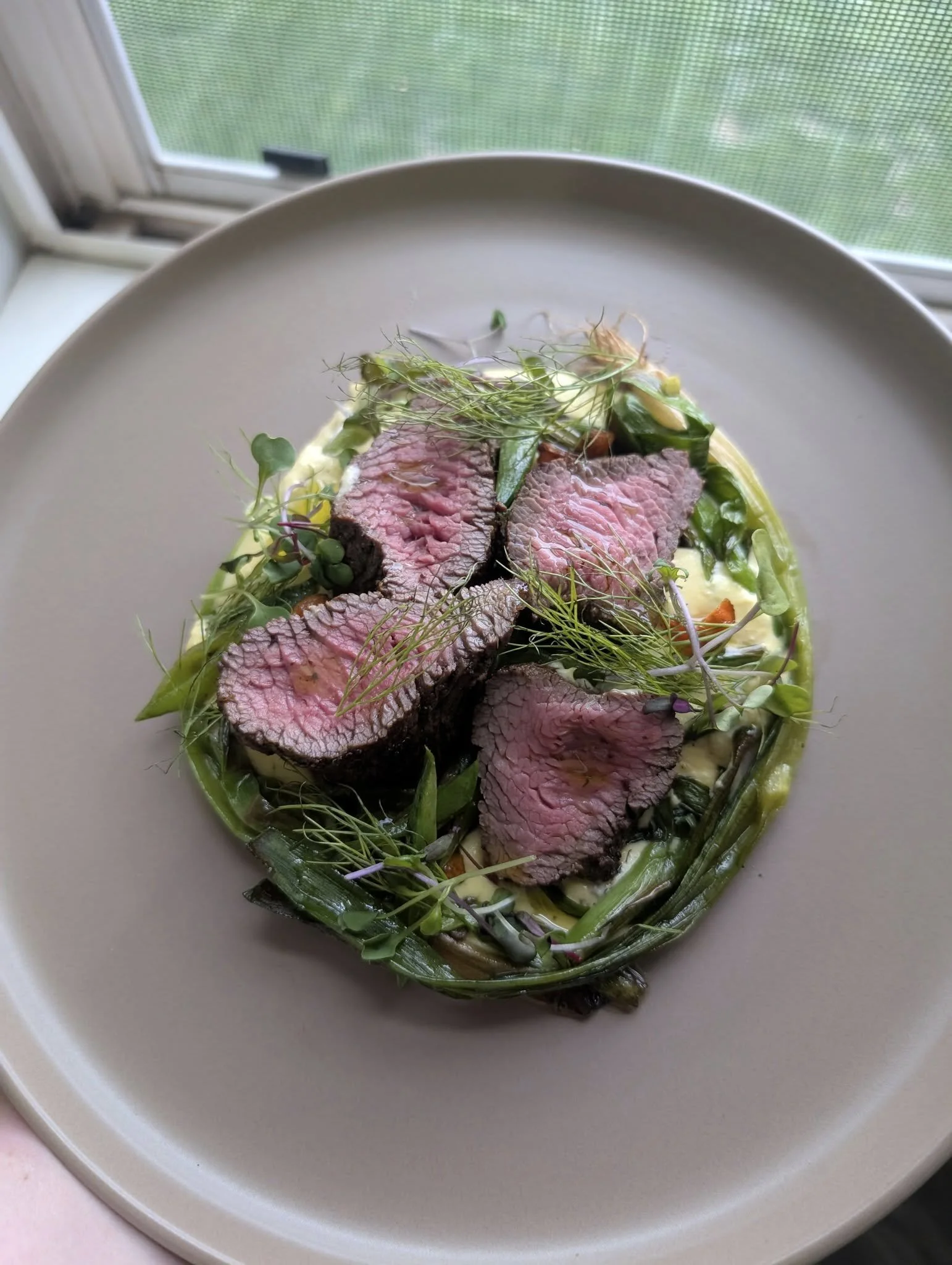 A plated dish with sliced medium-rare steak on top of greens and vegetables, garnished with microgreens, on a white plate near a window.