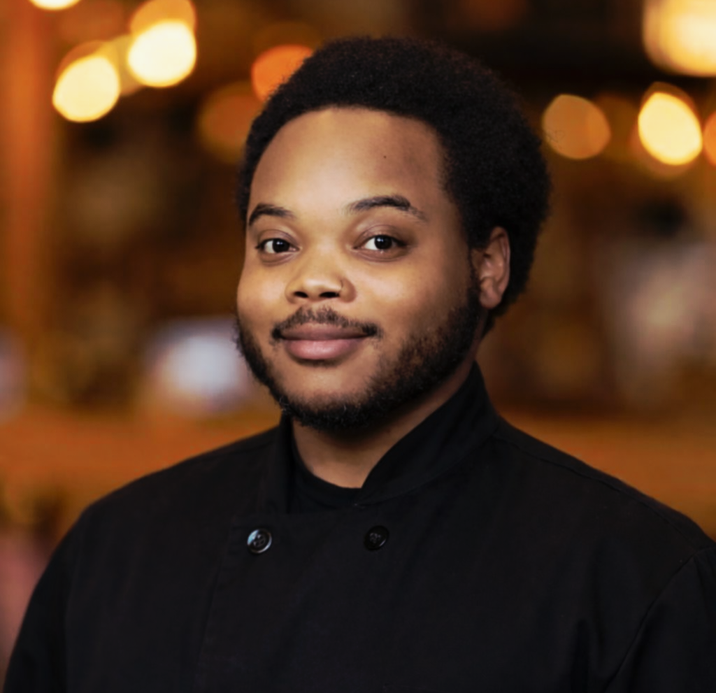 Portrait of a young man with a beard and afro hairstyle, wearing a black chef's coat, smiling in a warmly lit background.