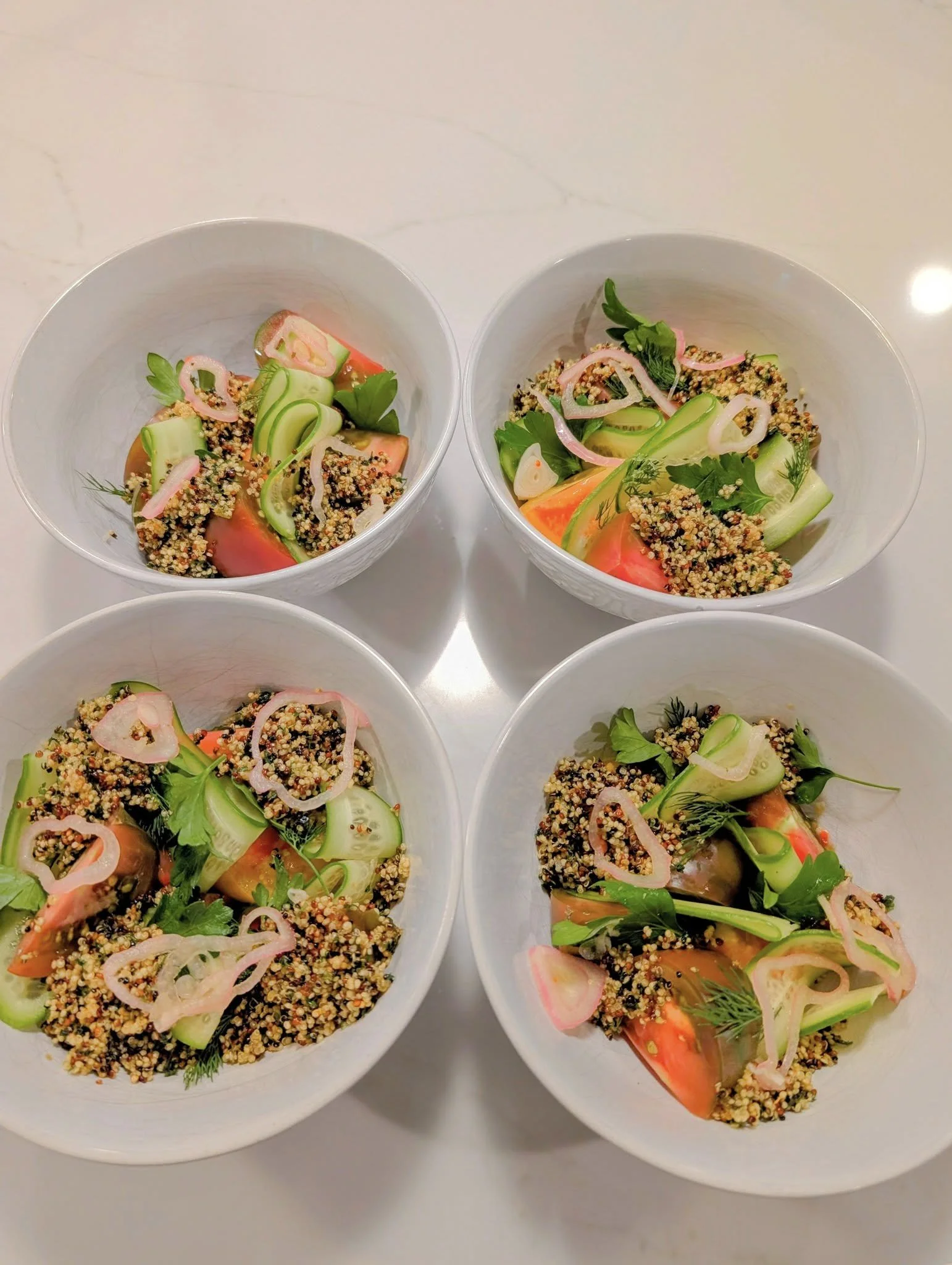 Four white bowls filled with salad containing quinoa, cucumber, tomato, herbs, and pickled onions, arranged on a white surface.