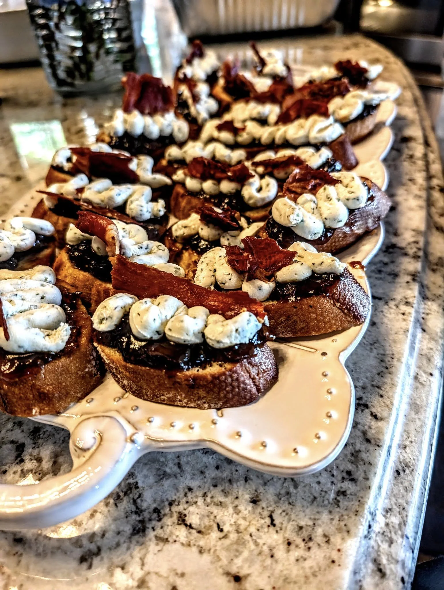 A decorative serving platter with multiple slices of toast topped with chocolate spread, whipped cream, bacon strips, and additional toppings, on a kitchen countertop.