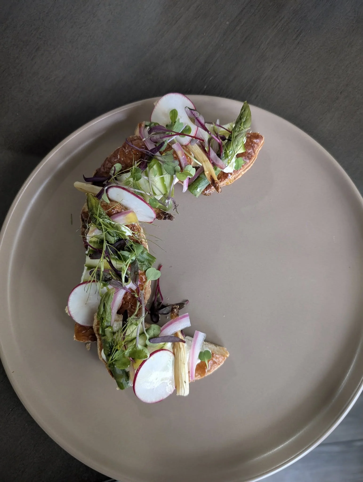 Plate with a crescent-shaped dish topped with microgreens, sliced radishes, and thinly sliced vegetables.