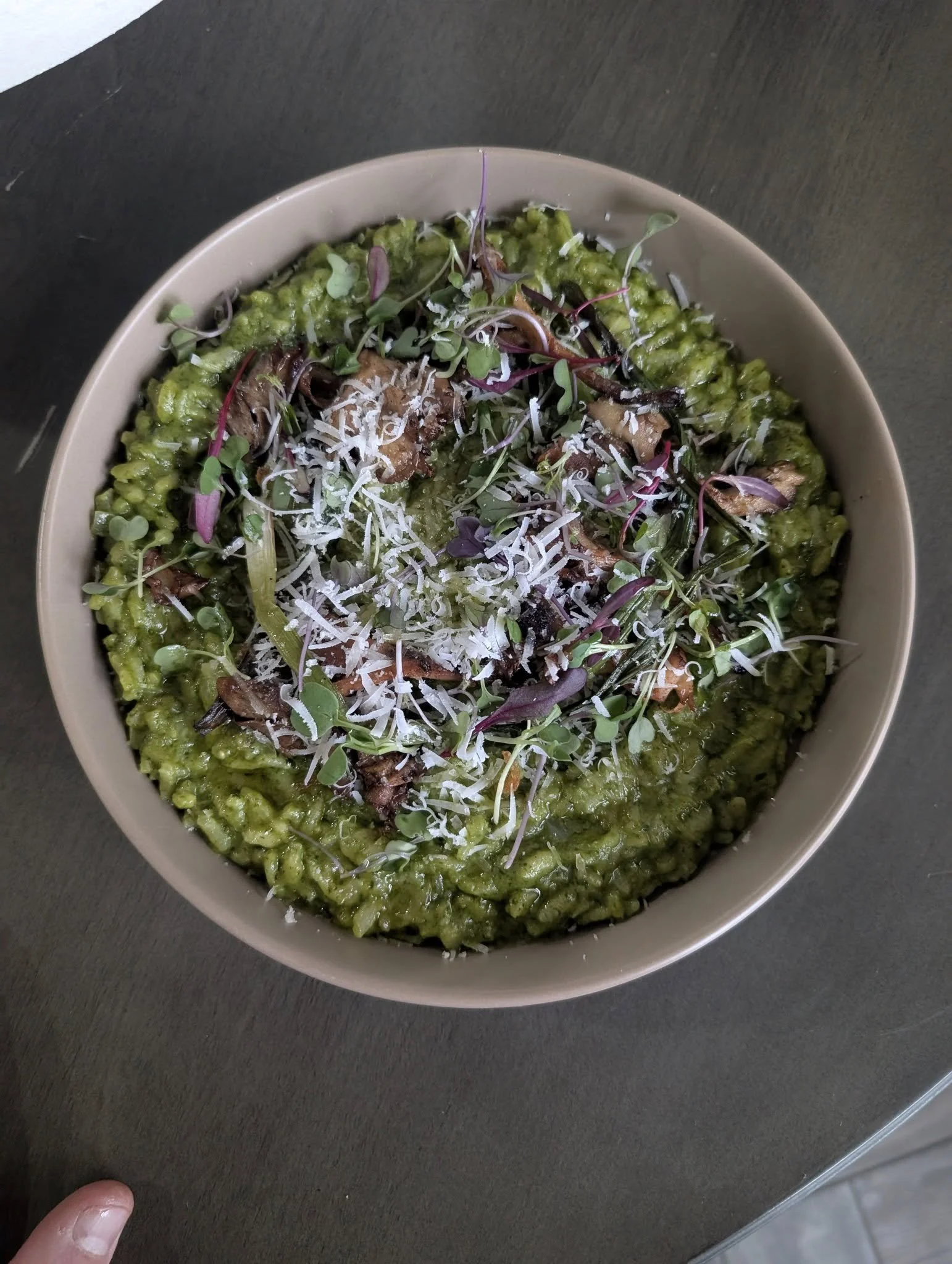 A bowl of green risotto topped with shredded cheese, microgreens, and mushrooms, placed on a black surface.