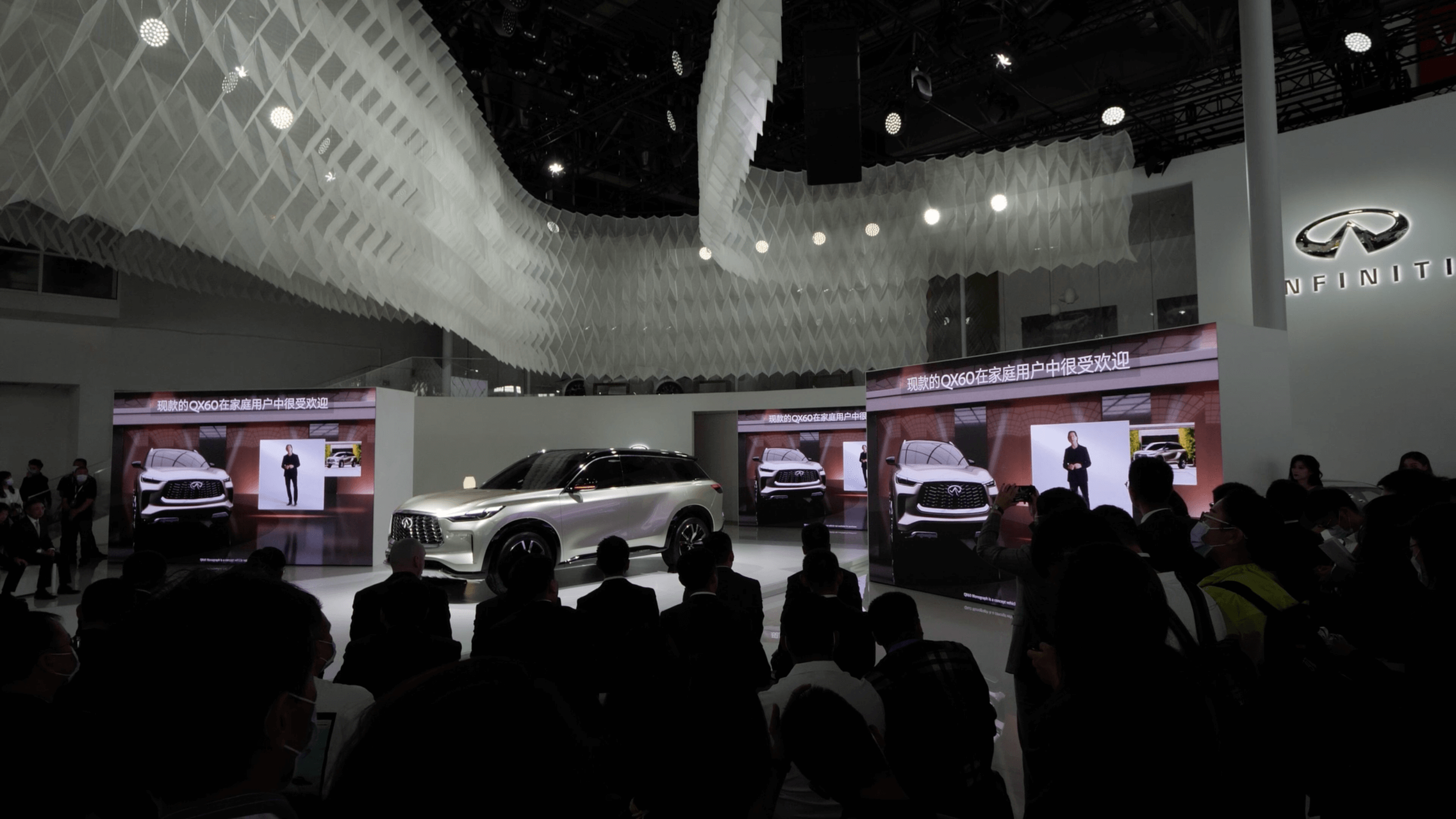 An automotive exhibition features several Infiniti cars on display with screens showing presenters and promotional content. The ceiling has a modern architectural design with white geometric panels and spotlights, and a crowd is gathered around the display.