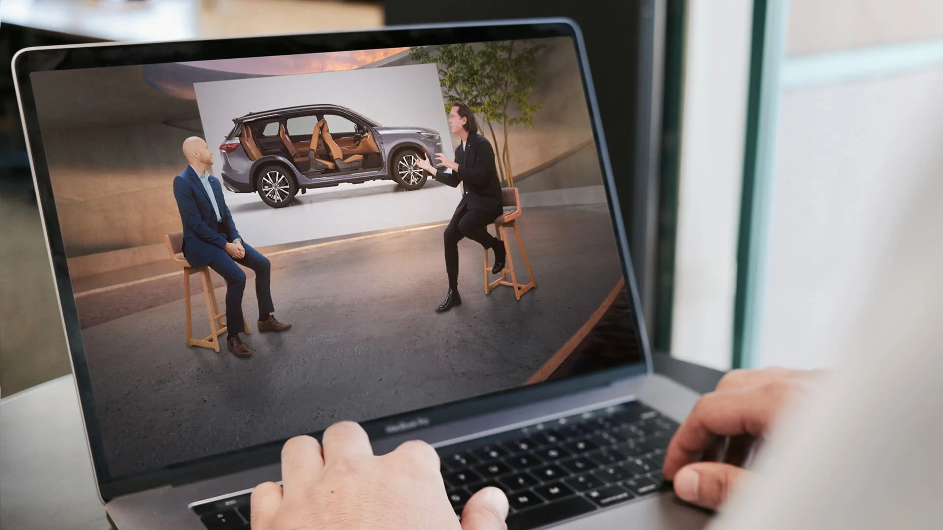 A person is working on a laptop, viewing a virtual interview or presentation of two women discussing a new car model with interior features. One woman is sitting on a stool, and the other is gesturing in front of a large screen showing the car.