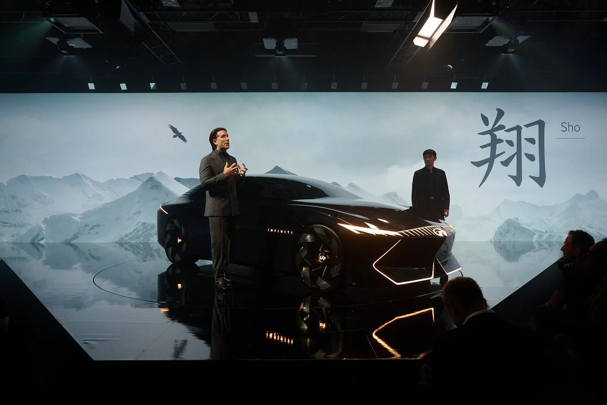 A man and a woman are standing next to a sleek black concept car on a stage with a mountain landscape backdrop and Chinese characters, including the word 'Sho', displayed behind them.