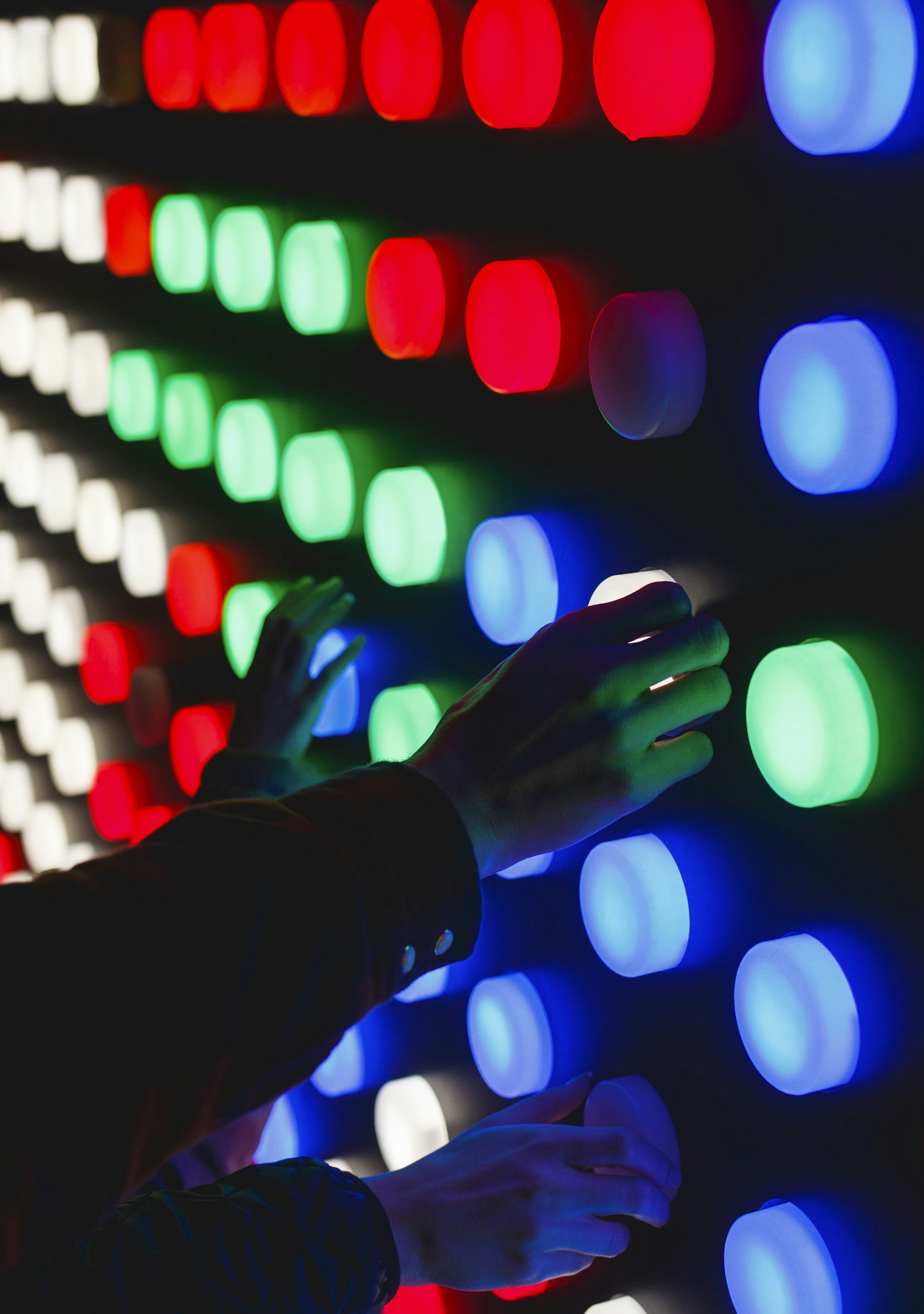 A person touching a wall of colorful LED lights with their hand, with the lights arranged in diagonal rows of red, white, green, and blue colors.