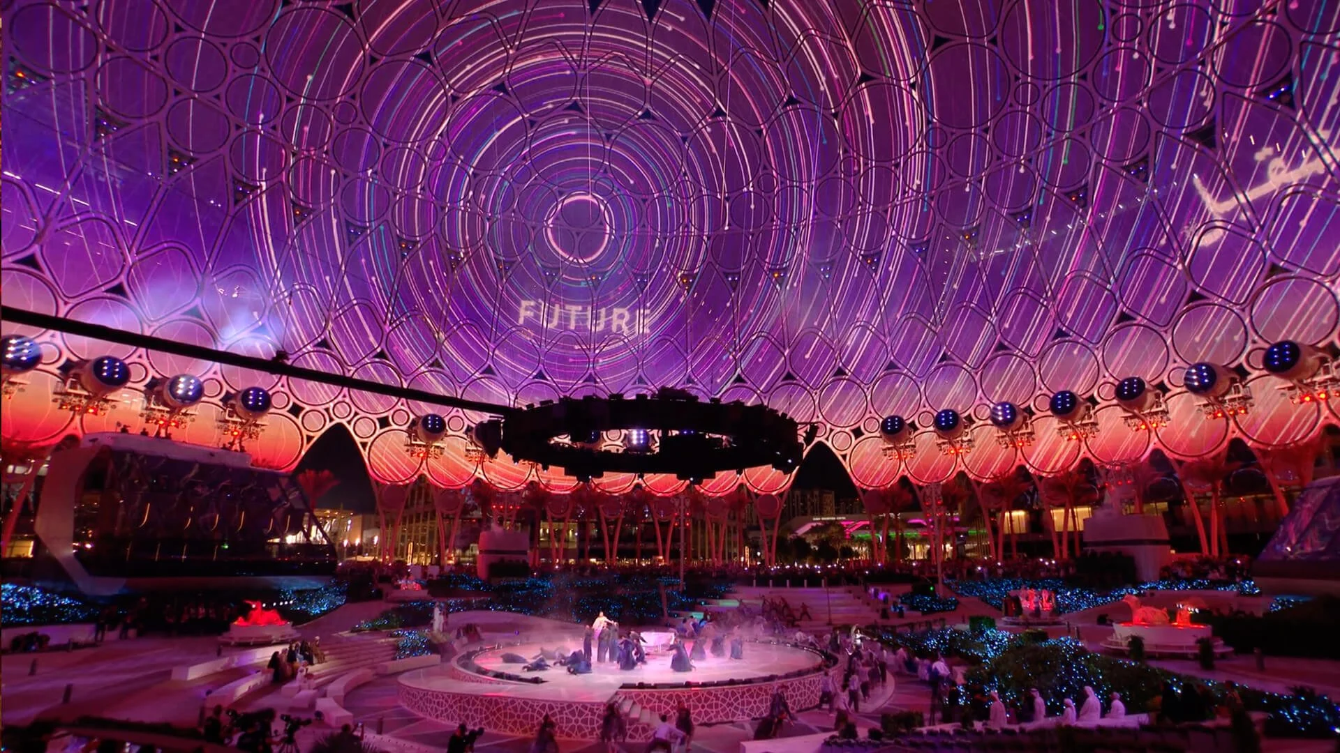 A large, illuminated domed structure with a pattern of interconnected circles and concentric circles, featuring colorful neon lights in pink, purple, and blue hues, with people sitting and walking around at sunset.