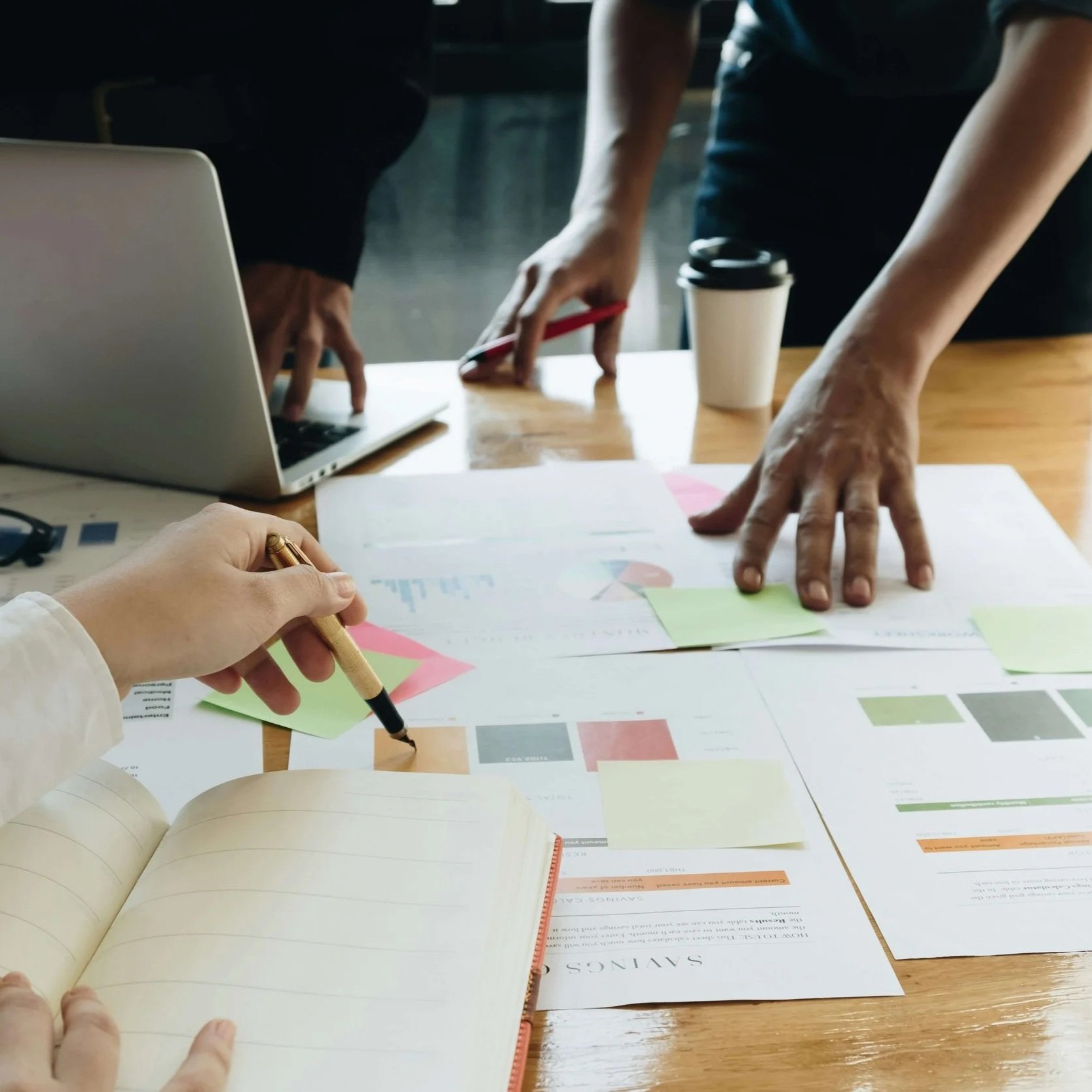 People working at a desk with papers, charts, a laptop, and a coffee cup, engaging in a collaborative discussion.