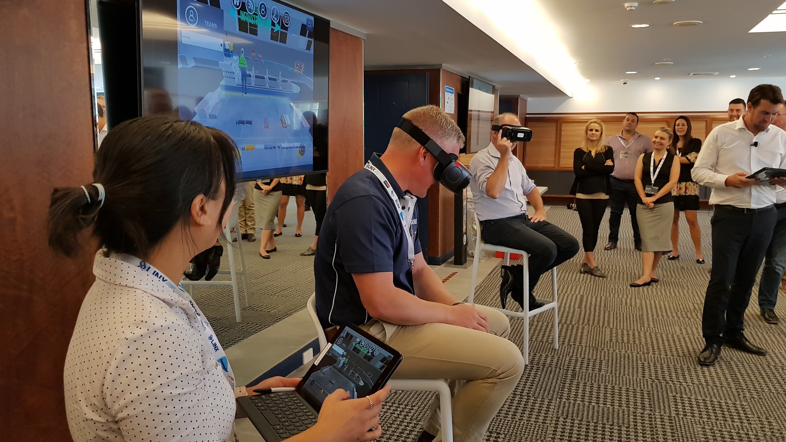 A group of people at a technology exhibition or conference. One person is wearing a virtual reality headset, and others are watching or participating. A woman in the foreground is working on a tablet. A large screen displays a digital interface. People are engaged and observing the presentation.