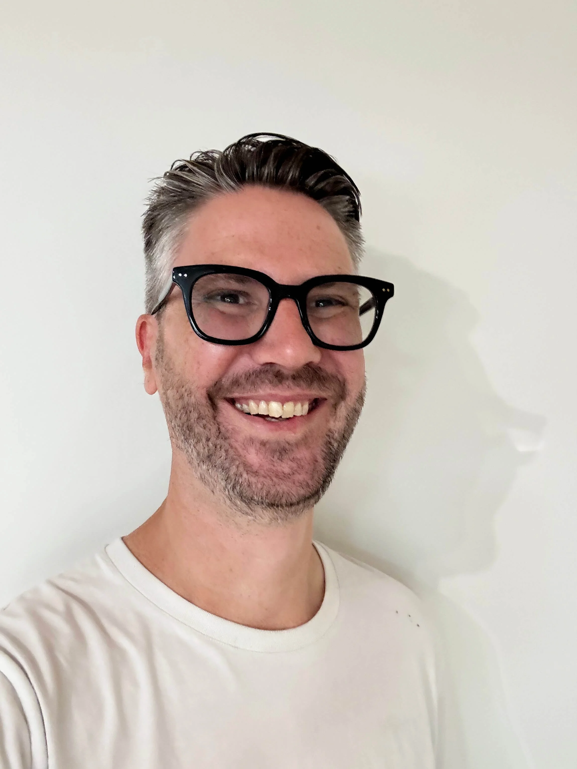 A smiling man with short dark hair, wearing large black glasses and a white T-shirt, standing against a plain light-colored wall.