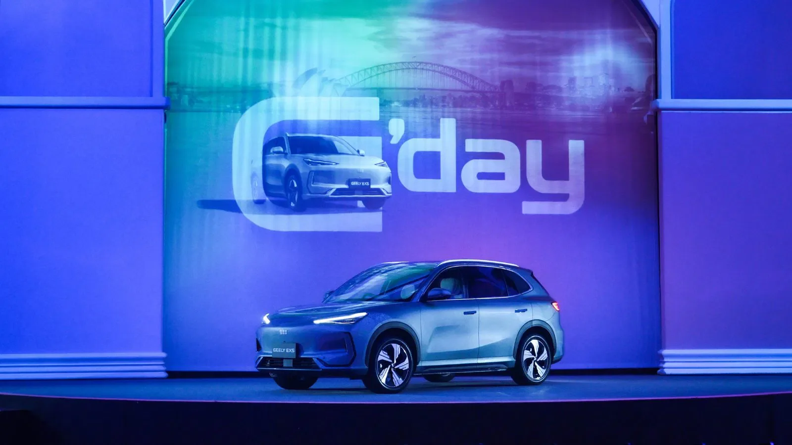 A silver electric compact SUV on stage at a car show for the Geely Australia Launch, with a large screen behind displaying the logo for 'G’day' and an image of the same vehicle.