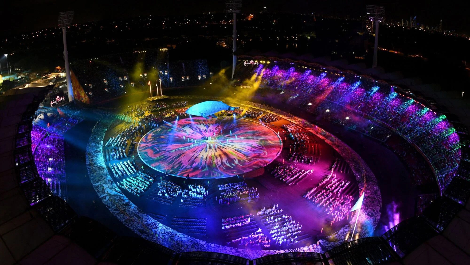 Nighttime view of the Commonwealth Games Opening Ceremony 2018 with vibrant, colorful lights and a whale-shaped structure in the center, surrounded by an illuminated track and audience seating.