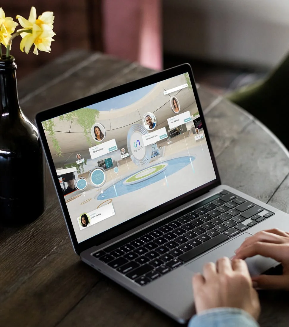 Person using a MacBook laptop on a wooden table, displaying a virtual social network or conference interface with profile pictures and connections, next to a black vase with yellow flowers.