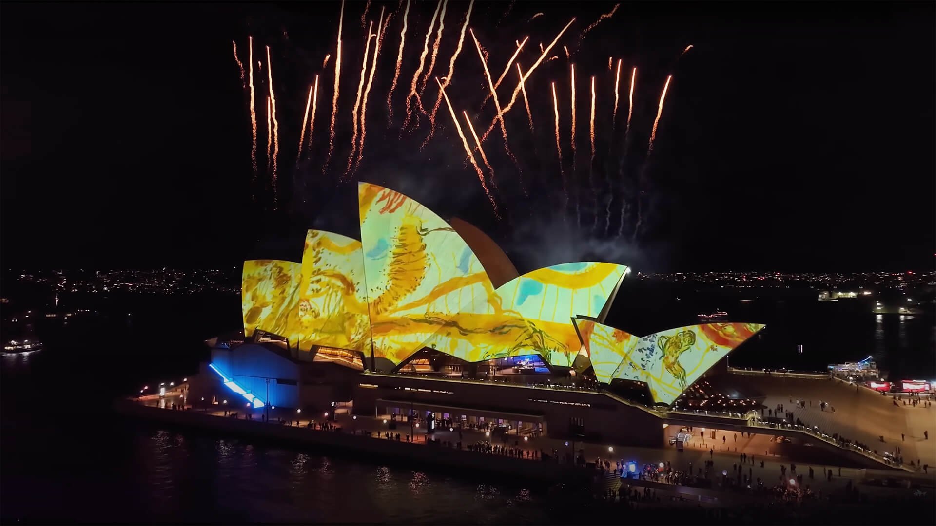 Night view of the Sydney Opera House illuminated with colorful projection, with fireworks displaying overhead in the night sky.