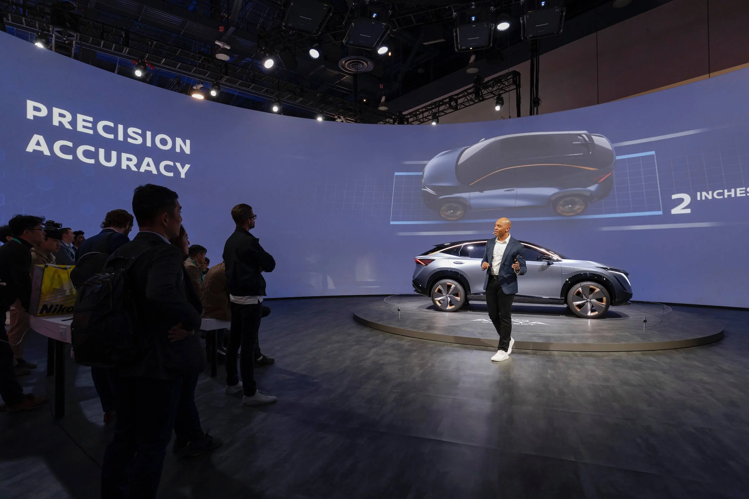At a tech conference, a presenter stands on a stage in front of a large curved screen displaying a futuristic vehicle design. The screen shows the words 'Precision' and 'Accuracy,' along with a digital rendering of a sleek, autonomous car and measure