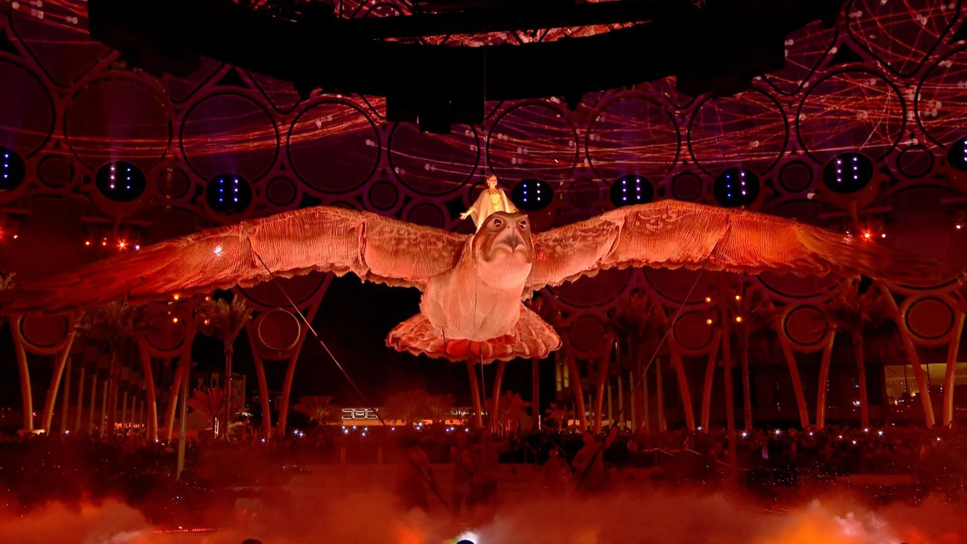 A large, illuminated butterfly sculpture with a person standing on its back, surrounded by a vibrant, colorful light display at night.