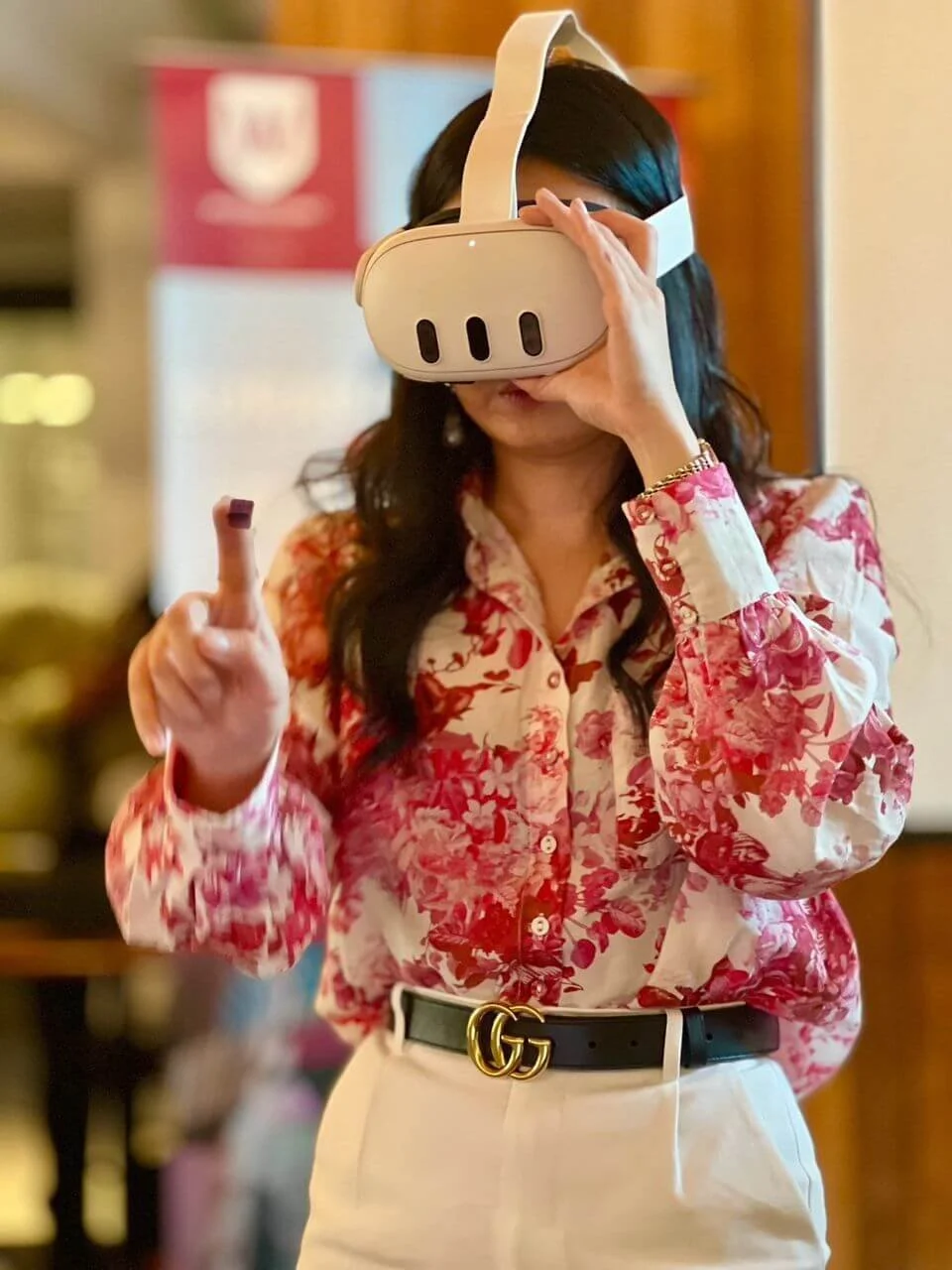 Woman wearing a floral blouse and white pants with a black Gucci belt, using a virtual reality headset in an indoor setting.