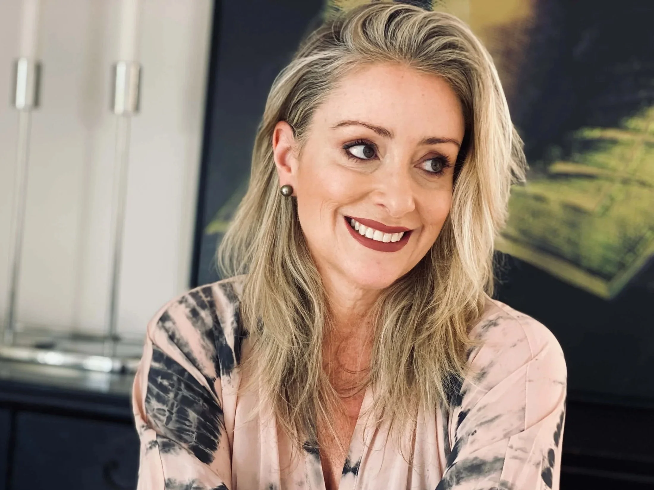 A woman with blonde hair, wearing pearl earrings and a light-colored blouse with a tie-dye pattern, smiling and looking to her side, with a blurred background showing a piece of framed artwork.