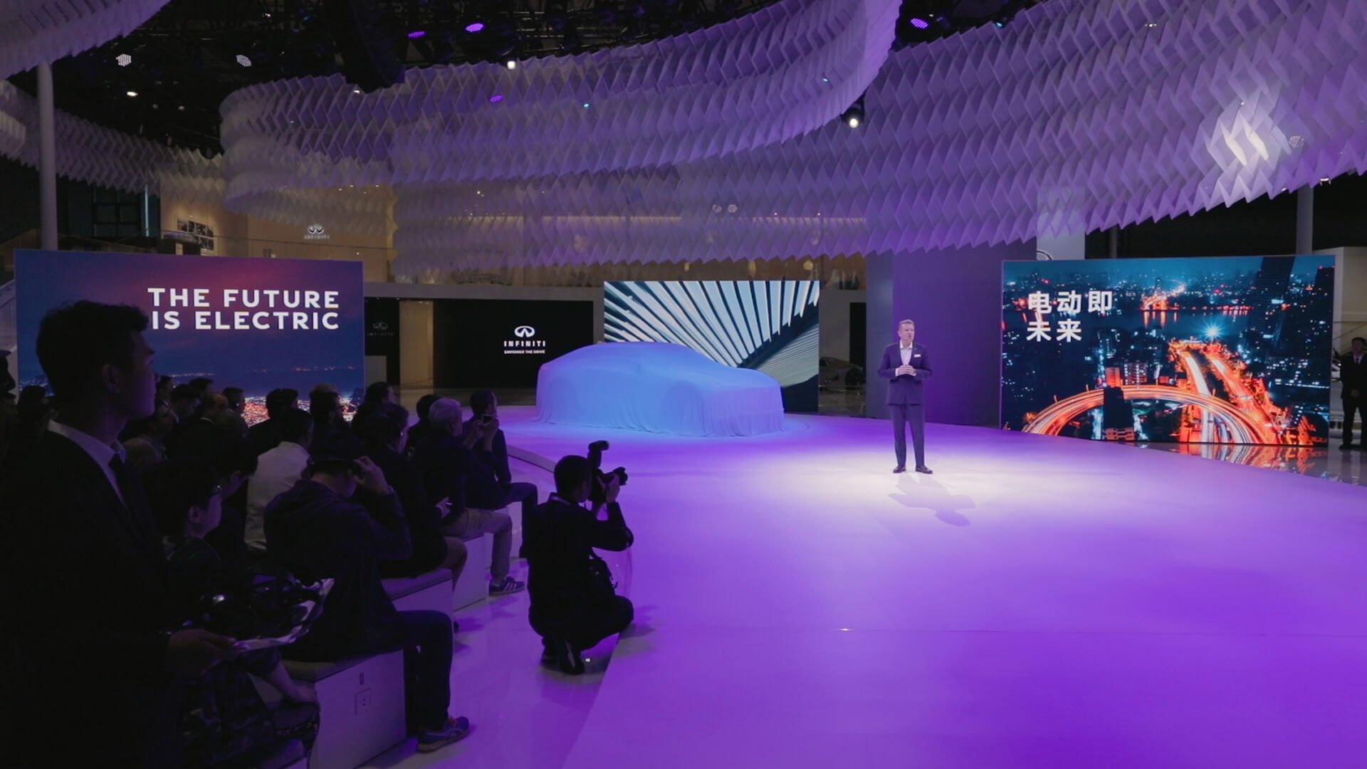 A man in a suit standing on stage at a presentation, with large LED screens displaying cityscape images and Chinese characters, an audience seated to the left, and a covered object in the center.
