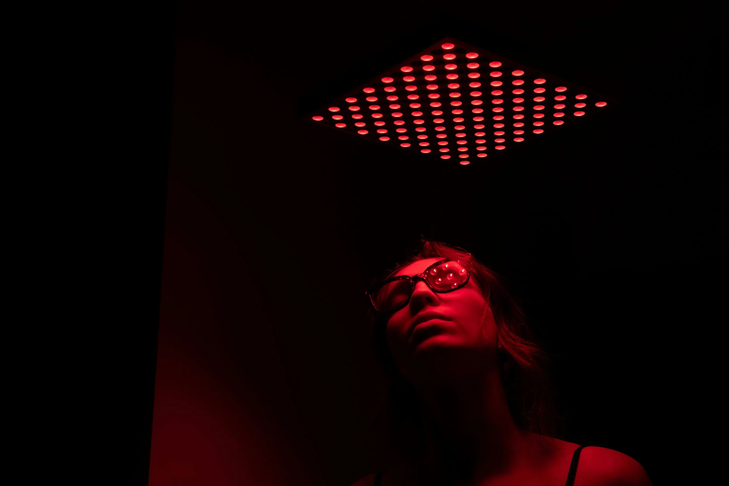 A woman with sunglasses looking up in a dark room illuminated by red light from a ceiling panel.