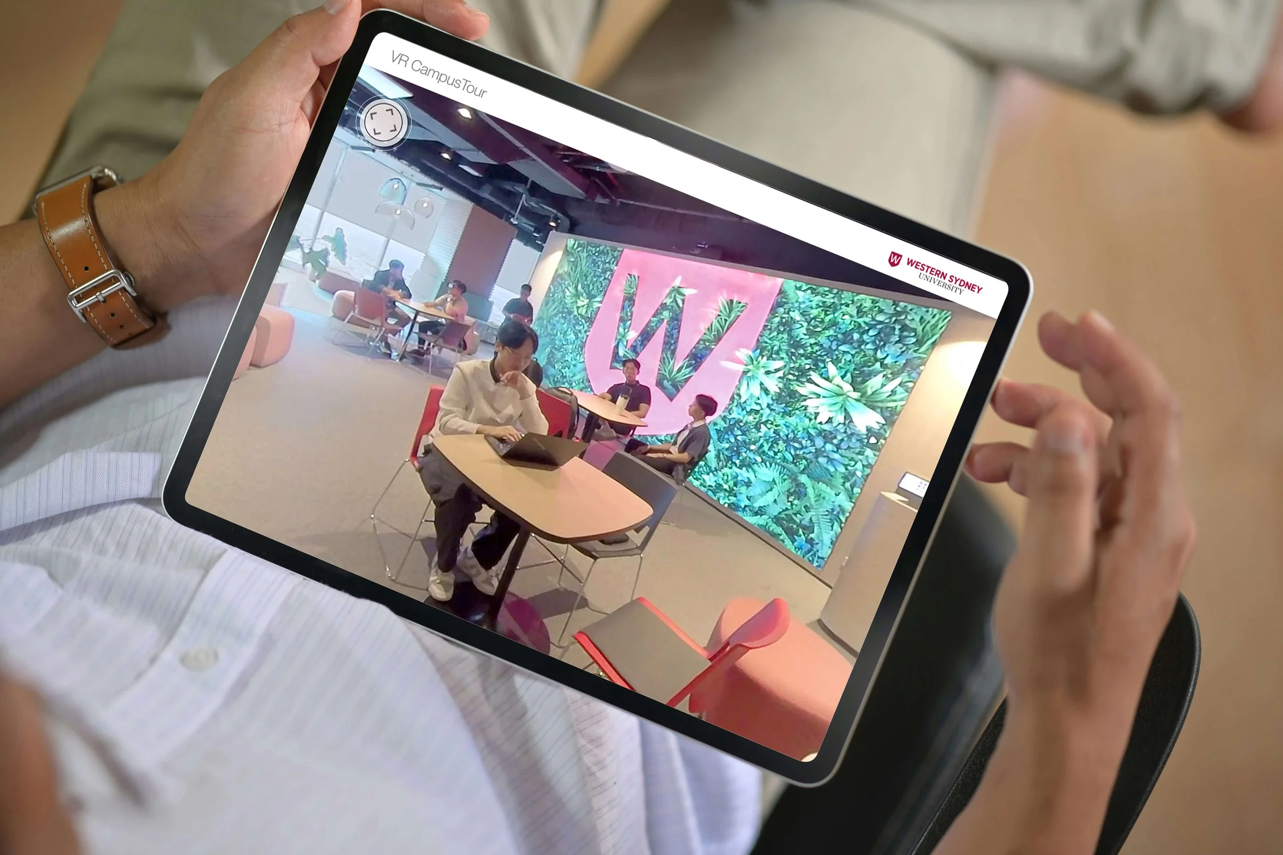 Person holding a tablet displaying a virtual tour of Western Sydney University, showing a lobby area with people sitting and a large digital screen with the university's logo and tropical-themed background.
