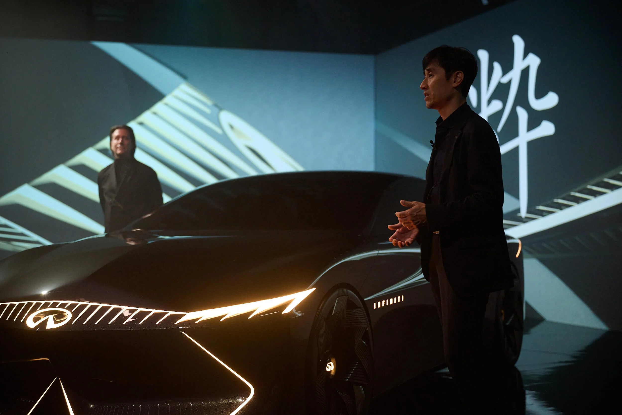 Two men in black suits standing next to a sleek, black Infiniti concept car with illuminated detailing, in front of a large illuminated backdrop with geometric patterns and words.