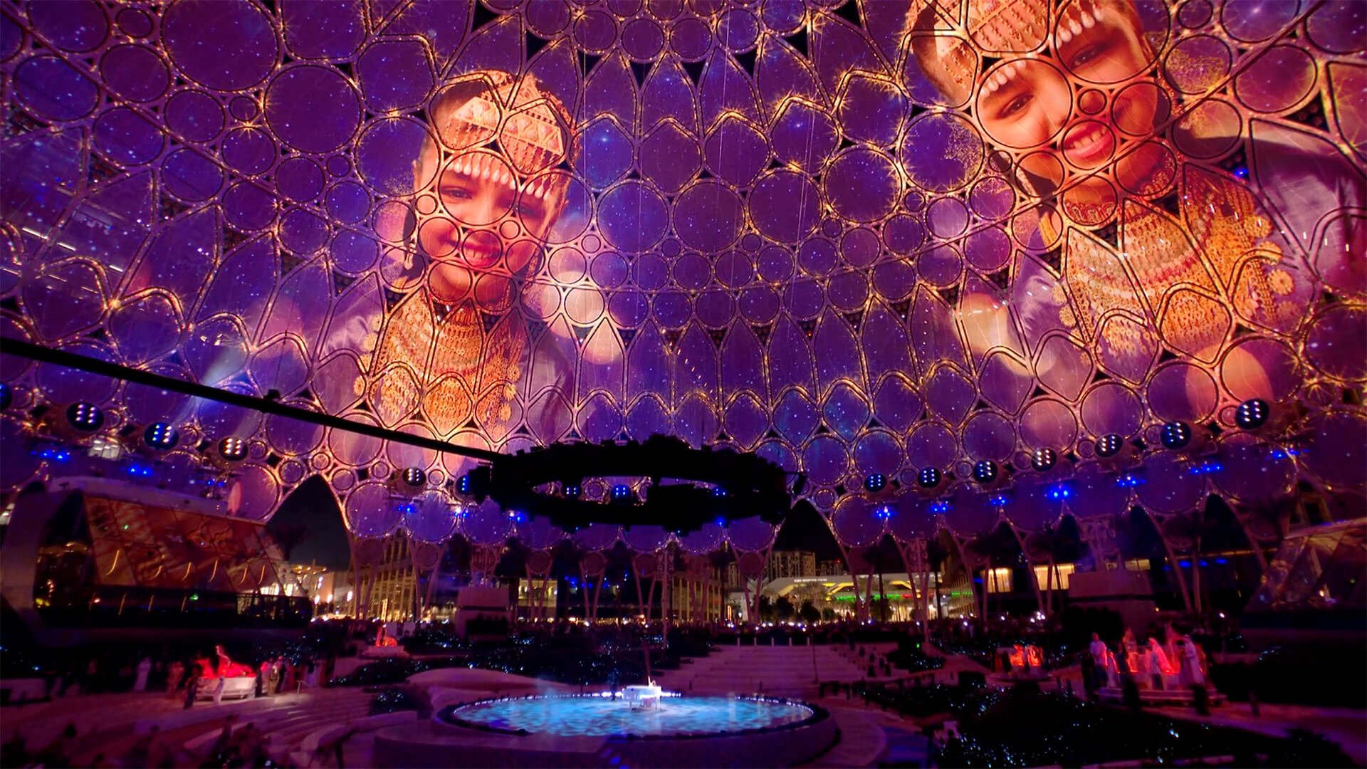 Large decorative dome structure with intricate geometric patterns, illuminated with purple and gold lighting, featuring images of smiling children on its surface, outside at night with people and vehicles below.