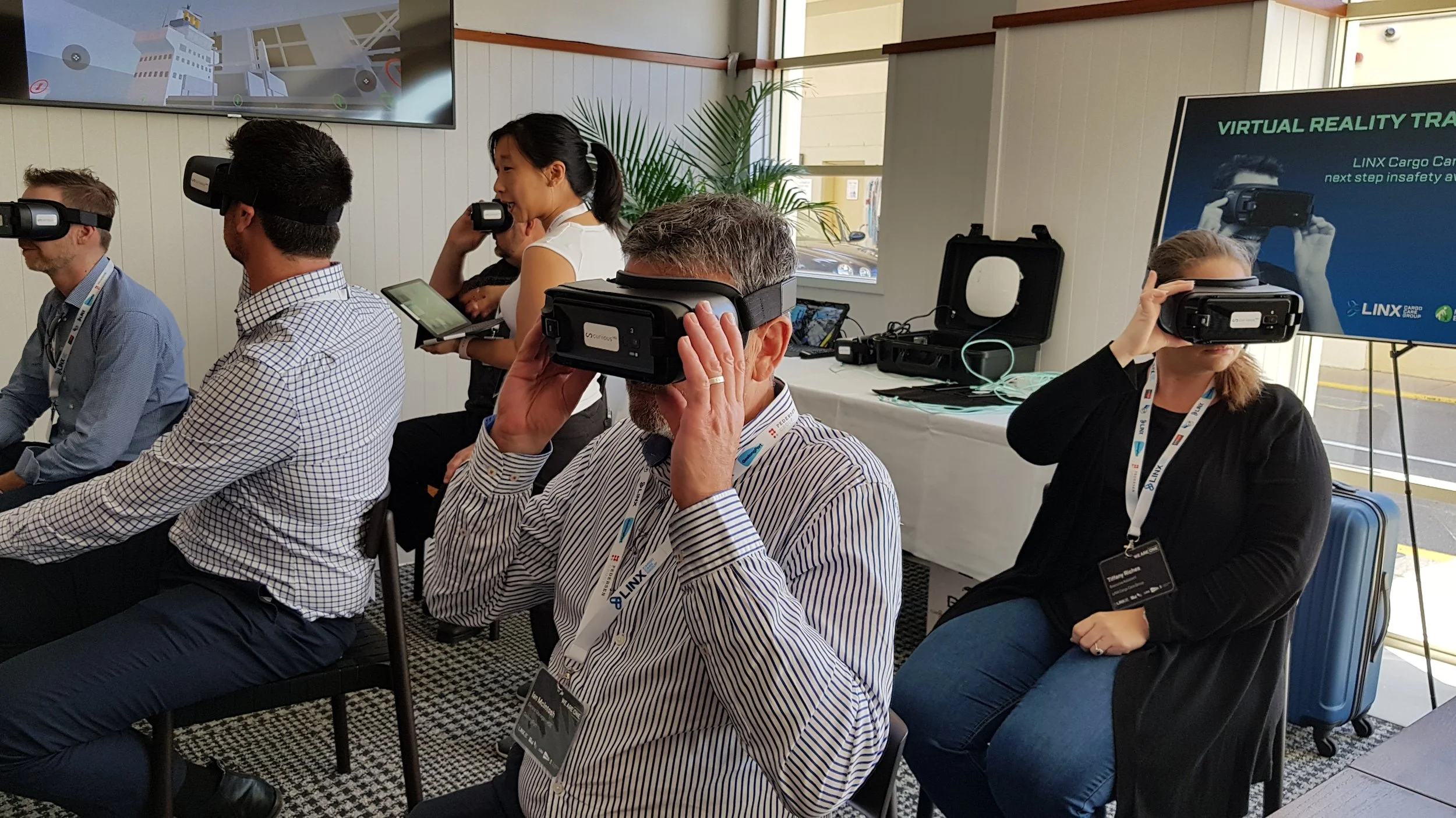 People wearing virtual reality headsets seated in a row during a presentation or event, with a large screen displaying VR-related content in the background.