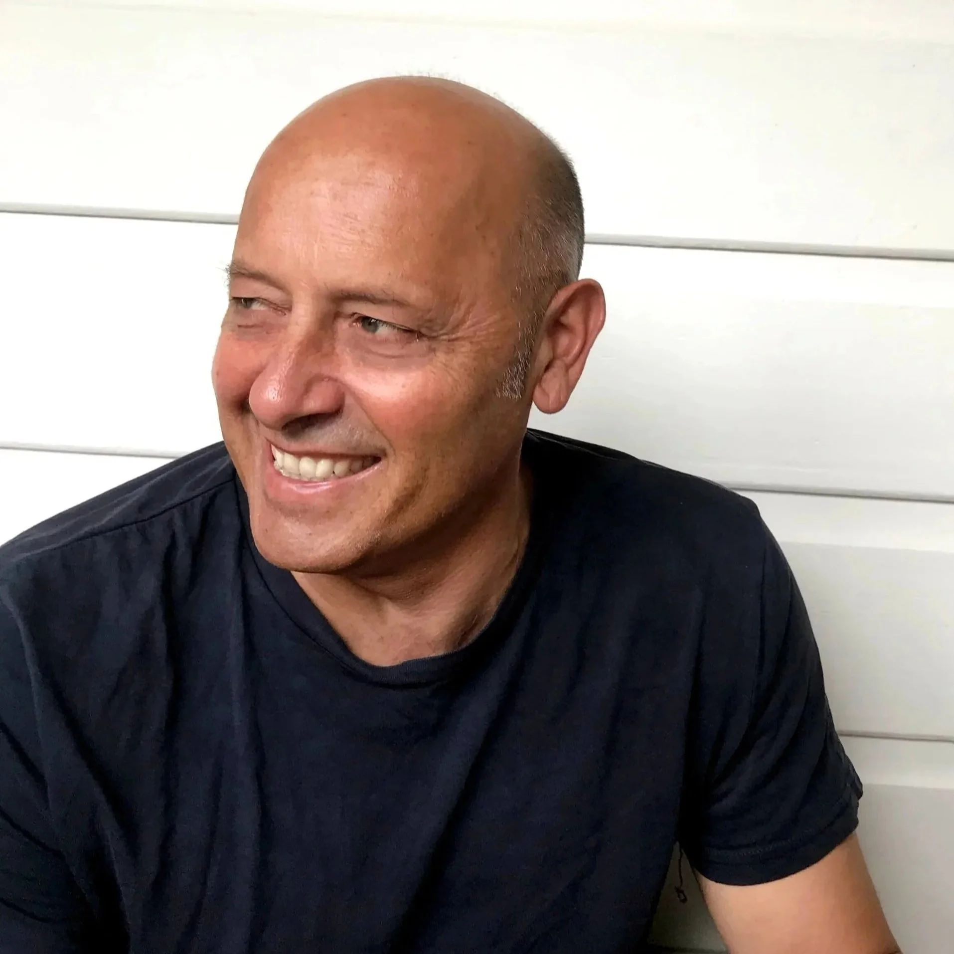 A smiling, bald man with a dark shirt, looking to the side, in front of a white wooden wall.