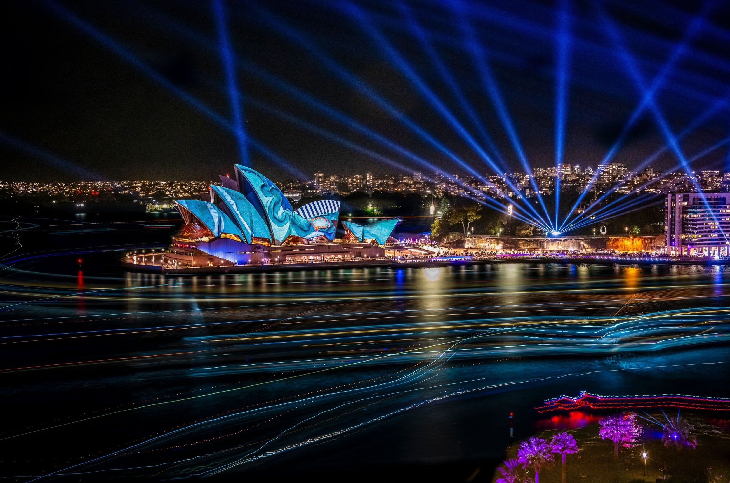 Night view of the Sydney Opera House illuminated with colorful lights, with blue laser beams shooting into the sky and city lights in the background.