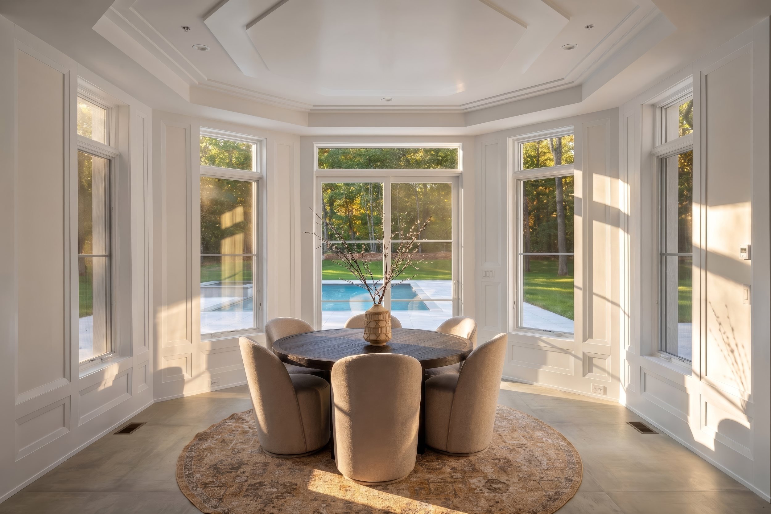 Round dining table in a bay-window dining room with coffered ceiling and pool view