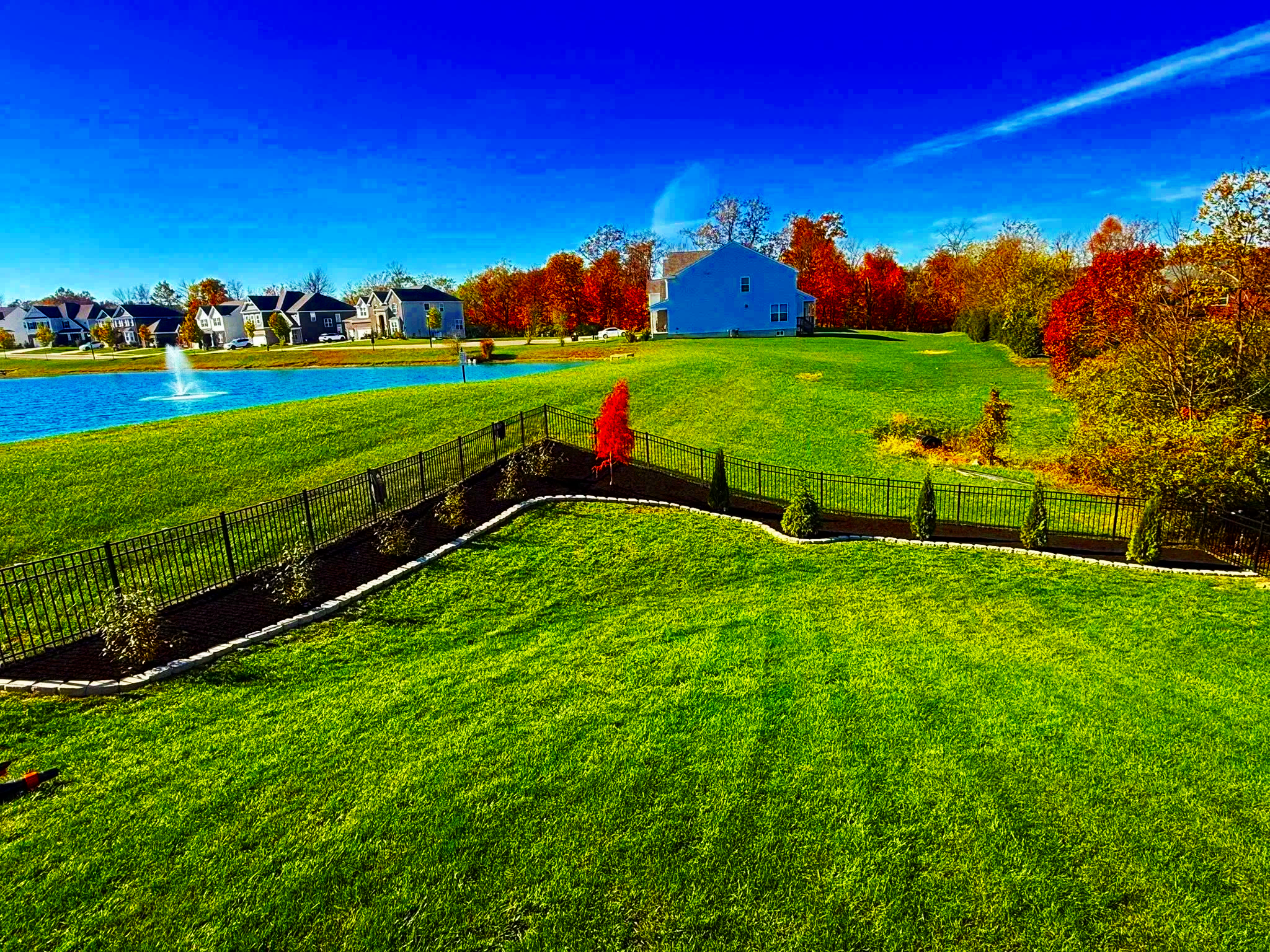 A scenic neighborhood with a pond featuring a fountain, houses, colorful trees, and well-maintained green lawn and landscaping under a bright blue sky.