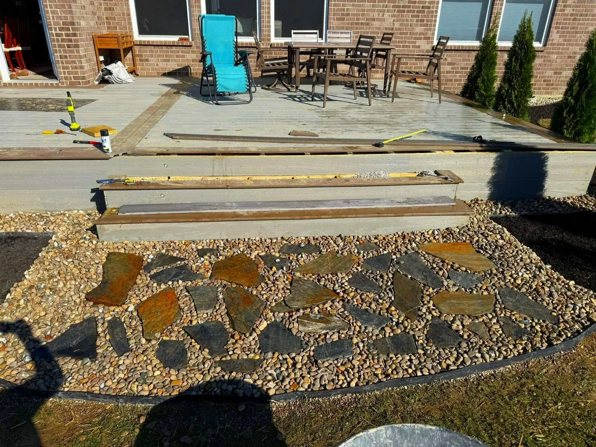 A backyard patio under construction with wooden steps, outdoor furniture, and a decorated stone pathway surrounded by small pebbles.