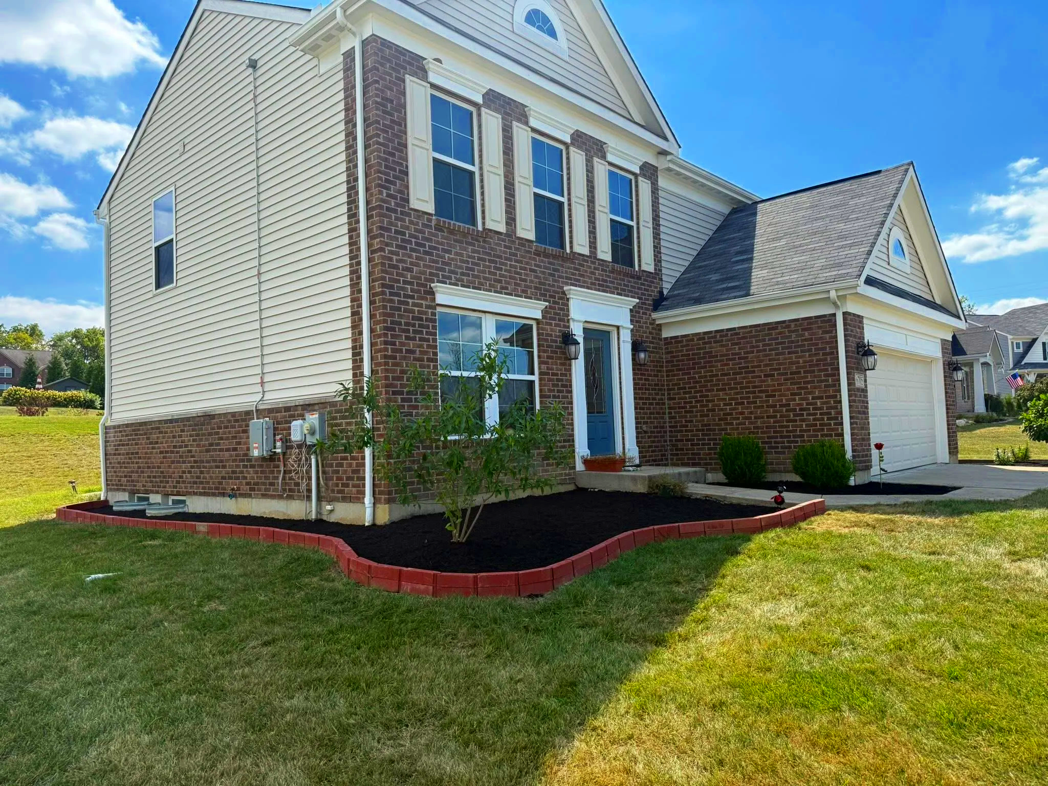 Front yard of a suburban house with a brick and siding exterior, a blue front door, and a single-car garage. The yard has freshly planted flower beds bordered with red bricks and a small shrub in the bed near the front door under a partly cloudy sky.