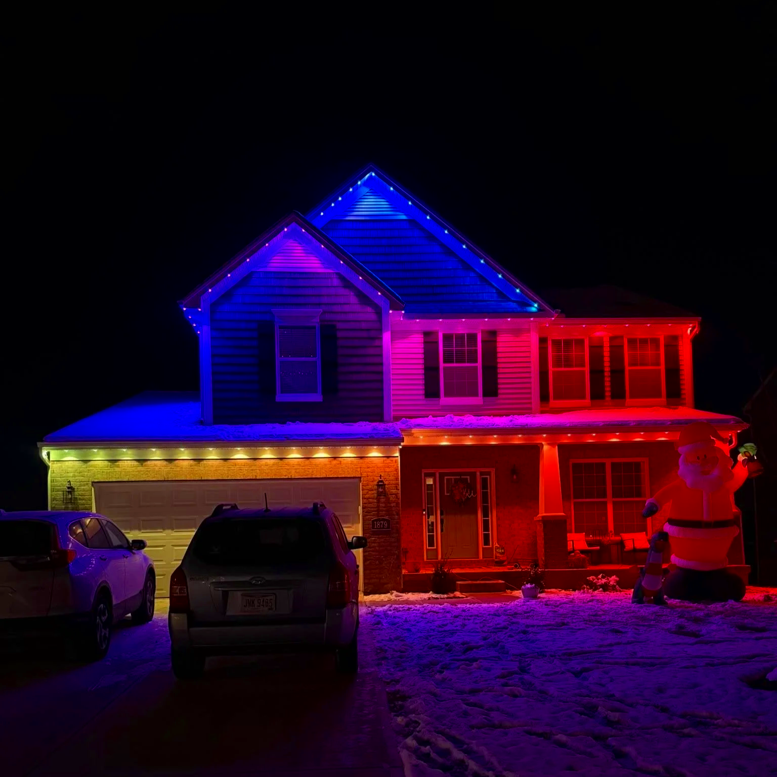 A two-story house decorated with multicolored Christmas lights at night, with snow on the ground and three parked cars in the driveway, including a Santa inflatable decoration outside.