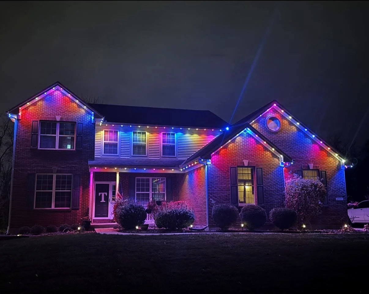 A two-story brick house decorated with colorful Christmas lights at night, with bushes and small lights in the front yard.