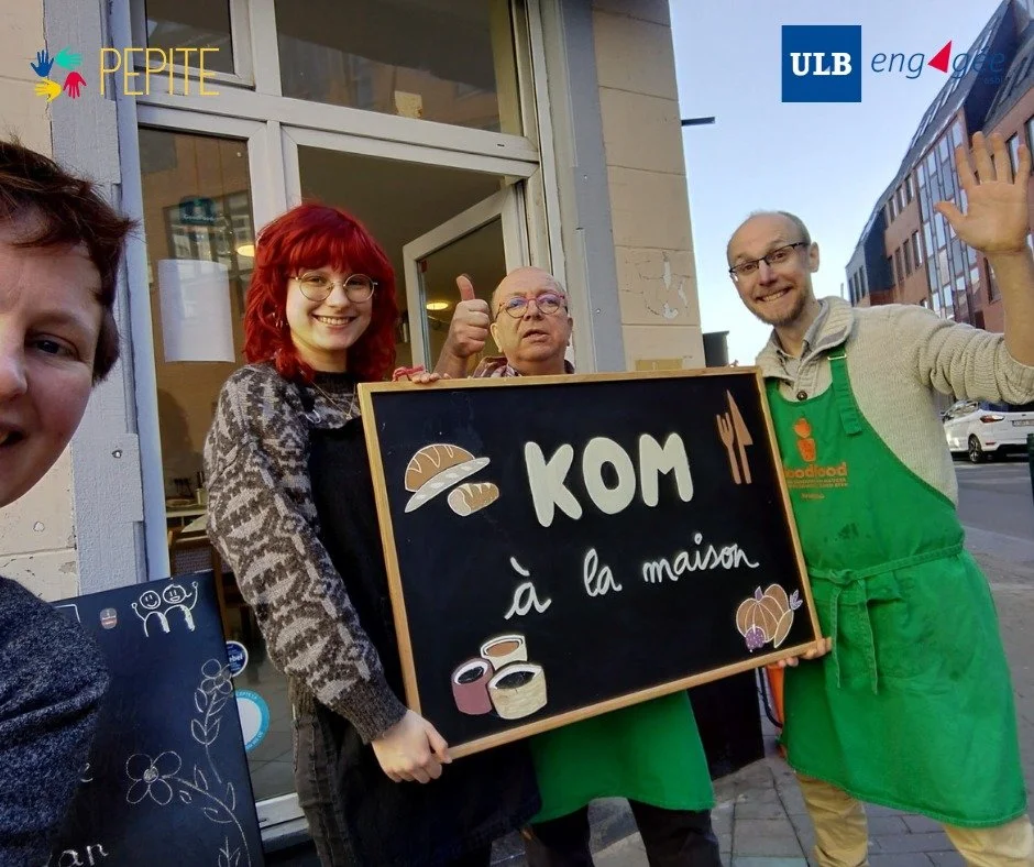 Deux hommes posent devant la devanture d'un restaurant appelé "KOM à la maison" avec une illustration dessiné de trois personnages portant des ustensiles de cuisine et un chef. La devanture est décorée avec des plantes en pot.