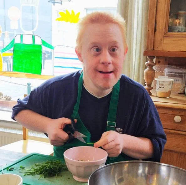 Un homme avec des cheveux blonds et roux, portant un tablier vert, cuisine dans une cuisine lumineuse, avec des herbes sur le plan de travail.