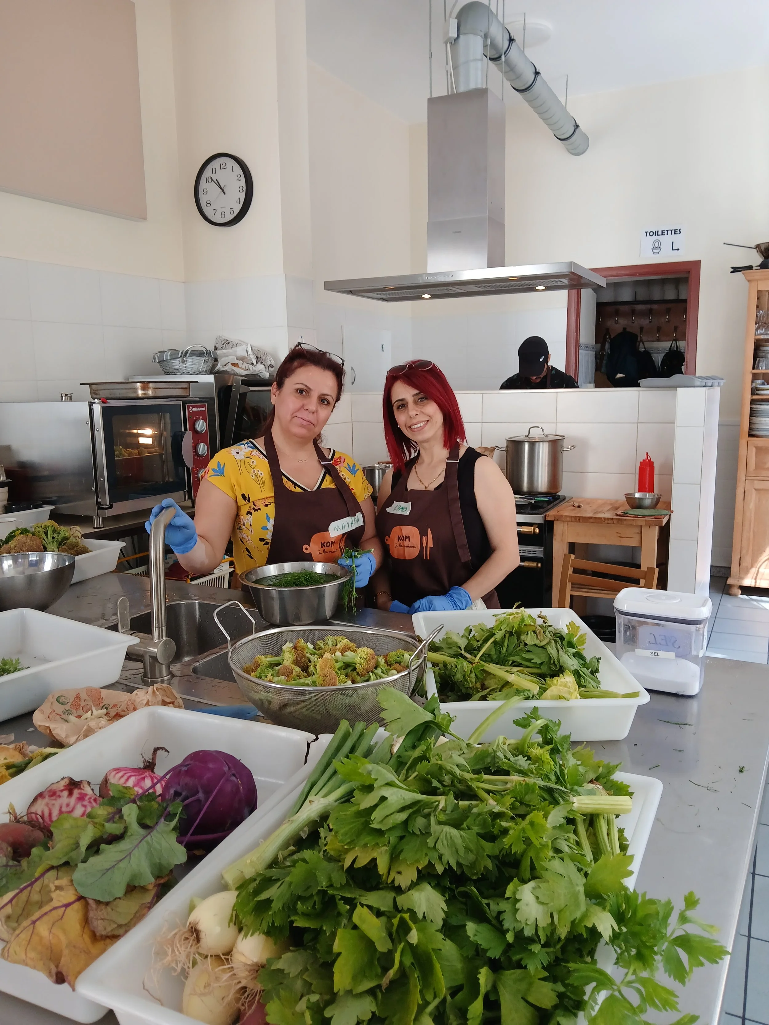 Deux femmes en tabliers et gants bleus dans une cuisine professionnelle, entourées de légumes frais et de plats préparés.