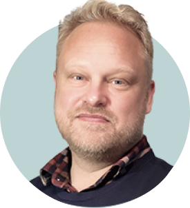 Portrait of a man with light-colored hair and beard, wearing a dark blazer over a checkered shirt, against a light background.